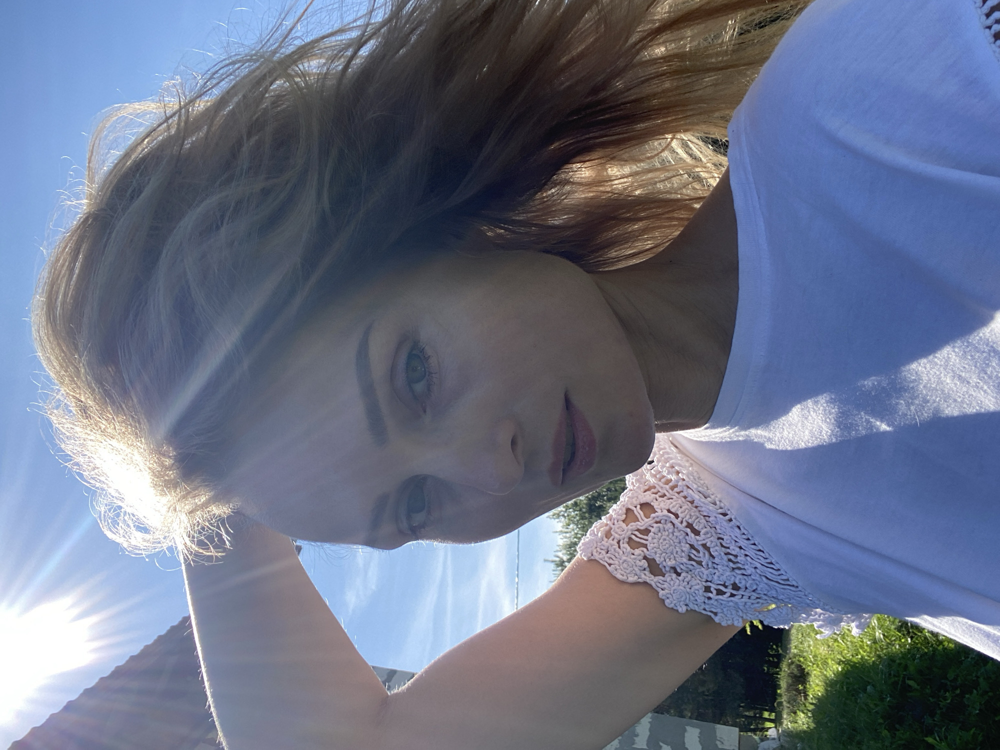 This is a close-up, outdoor selfie taken in the daytime. A woman with blonde hair is positioned in the foreground, with her right arm raised and hand in her hair. She is wearing a white t-shirt with intricate white crochet detailing on the sleeve. The sun is shining brightly from the upper left, creating lens flares and highlighting her hair. The background shows a clear blue sky, some greenery, and a portion of a building. The scene appears to be set in a rural or suburban environment, possibly a backyard or garden area. The location is identified as Міханавіцкі сельскі Савет, Belarus, suggesting a residential area outside of a larger town or city. The overall impression is one of a candid, sun-drenched moment.