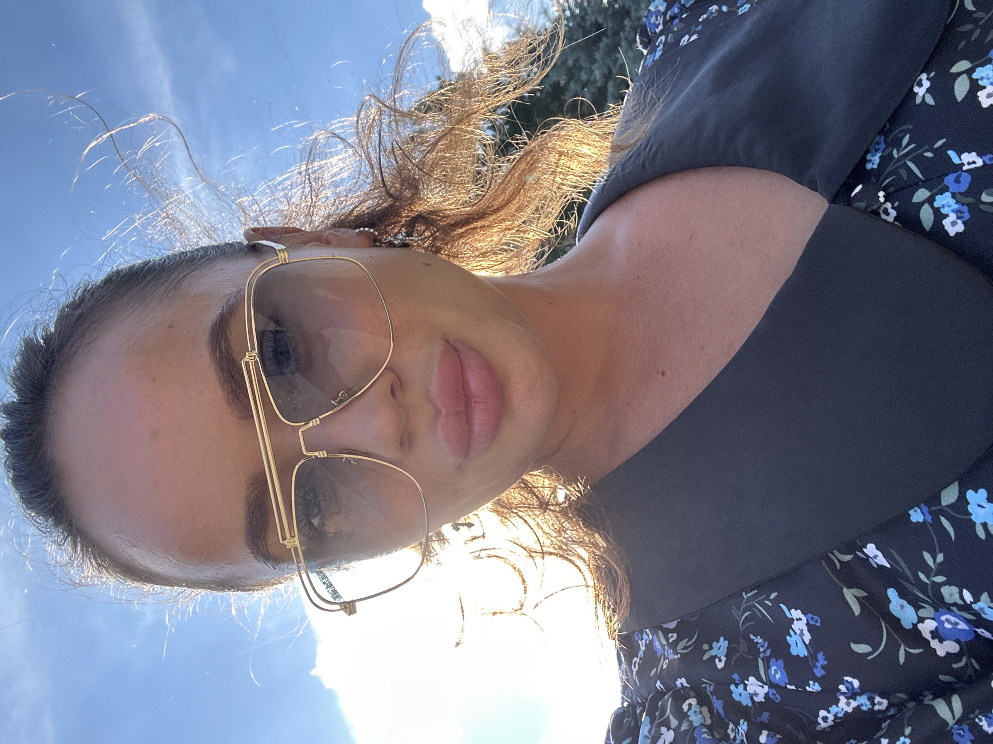 The original image is a close-up selfie of a woman outdoors, with a bright blue sky and some clouds in the background. The woman is wearing large, gold-rimmed aviator sunglasses and a dark dress with a blue floral pattern. Her hair is dark and appears windswept. The lighting suggests it is daytime, possibly late afternoon due to the warm glow on her hair. The background shows a glimpse of green foliage, possibly trees, and a bright light source, likely the sun, which is partially obscured by her head and sunglasses. There is no indication of the city of Duvankul', Russia, or any specific urban environment.
