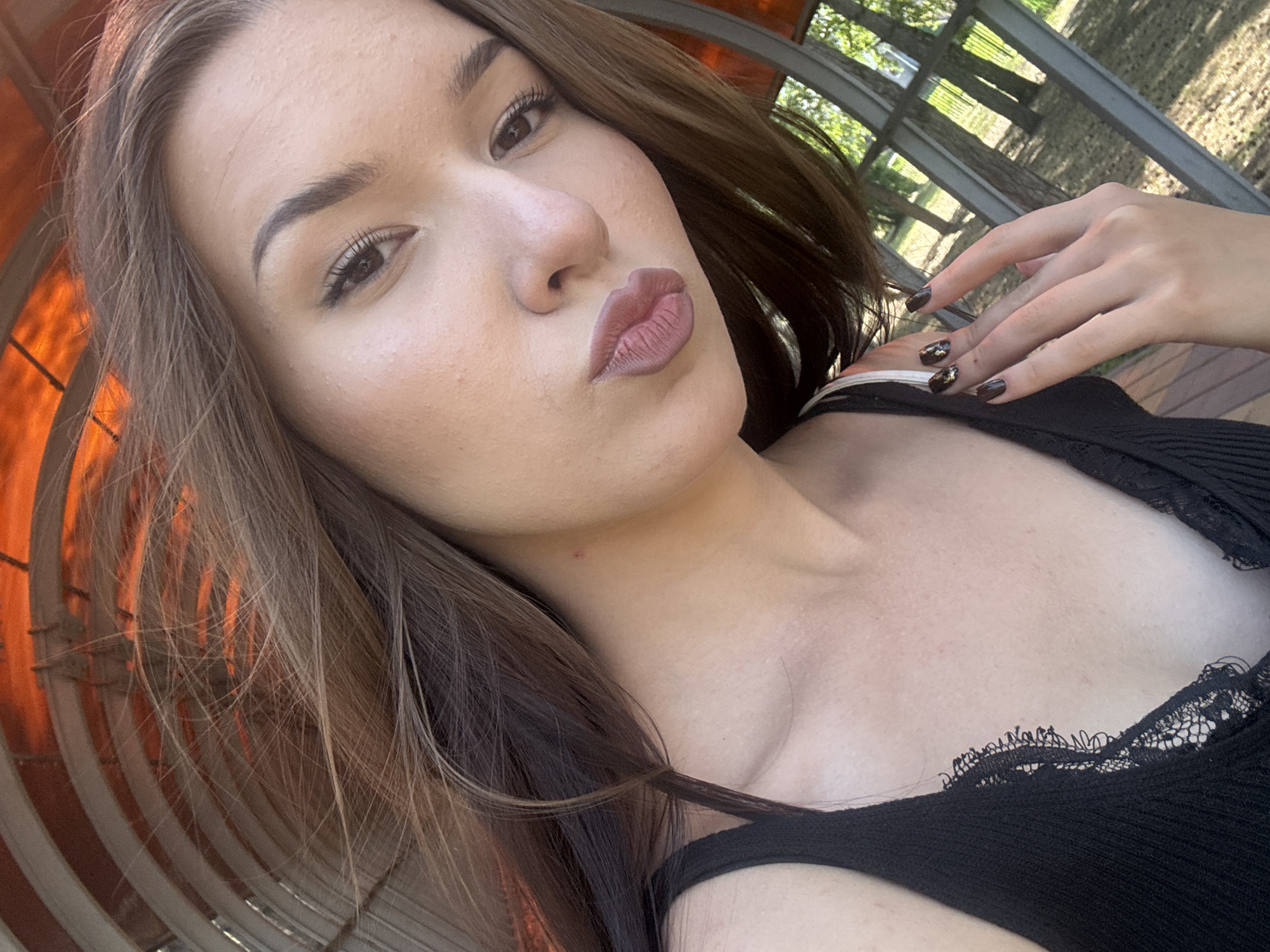 A young woman is taking a selfie outdoors. She has long, brown hair and is wearing a black tank top with lace trim. Her lips are pursed in a kiss. Her fingernails are painted dark brown with gold flecks. In the background, there are trees and a metal structure. The lighting suggests it is daytime. The image does not provide any information about a specific location in Belovo, Russia.