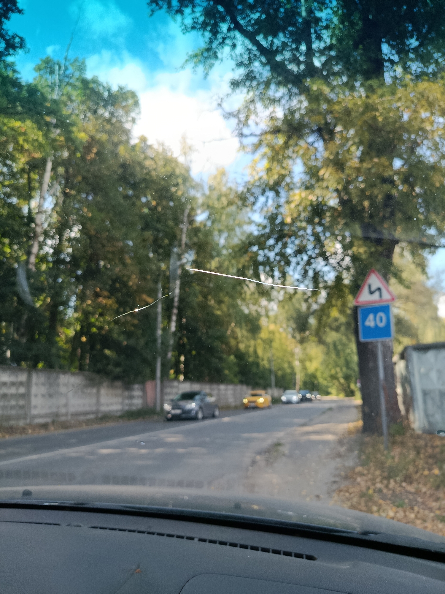 The image, taken from inside a vehicle, depicts a street scene in Orekhovo-Zuyevo, Russia. A cracked windshield is visible in the foreground, with reflections of trees and the sky. The road ahead is paved, with a concrete wall on the left side and trees lining the right side. Several cars are present on the road, and a traffic sign indicating a speed limit of 40 km/h is posted on the right. The overall environment suggests a sunny day with some foliage indicating an autumn setting.