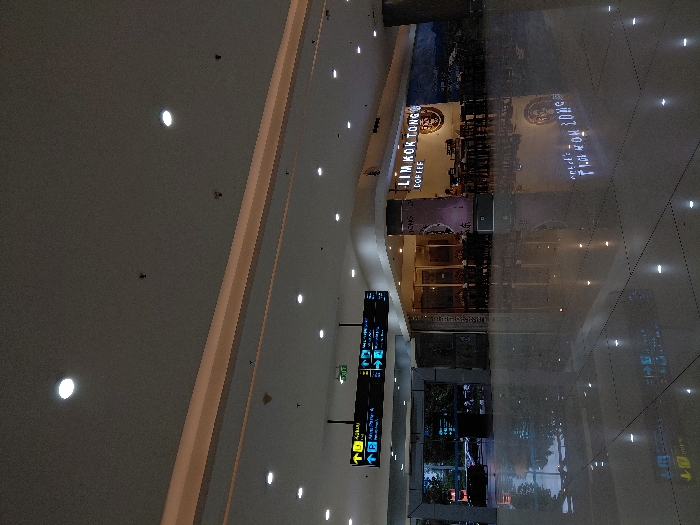 A brightly lit indoor space, possibly an airport or shopping mall, features a coffee shop named "Lim Kok Tong." The image reflects onto the polished floor, creating a mirrored effect. A large directional sign with arrows and symbols is present, guiding travelers or shoppers. The ceiling is adorned with evenly spaced recessed lights. A glimpse of the outside is visible through glass windows, hinting at trees or greenery.