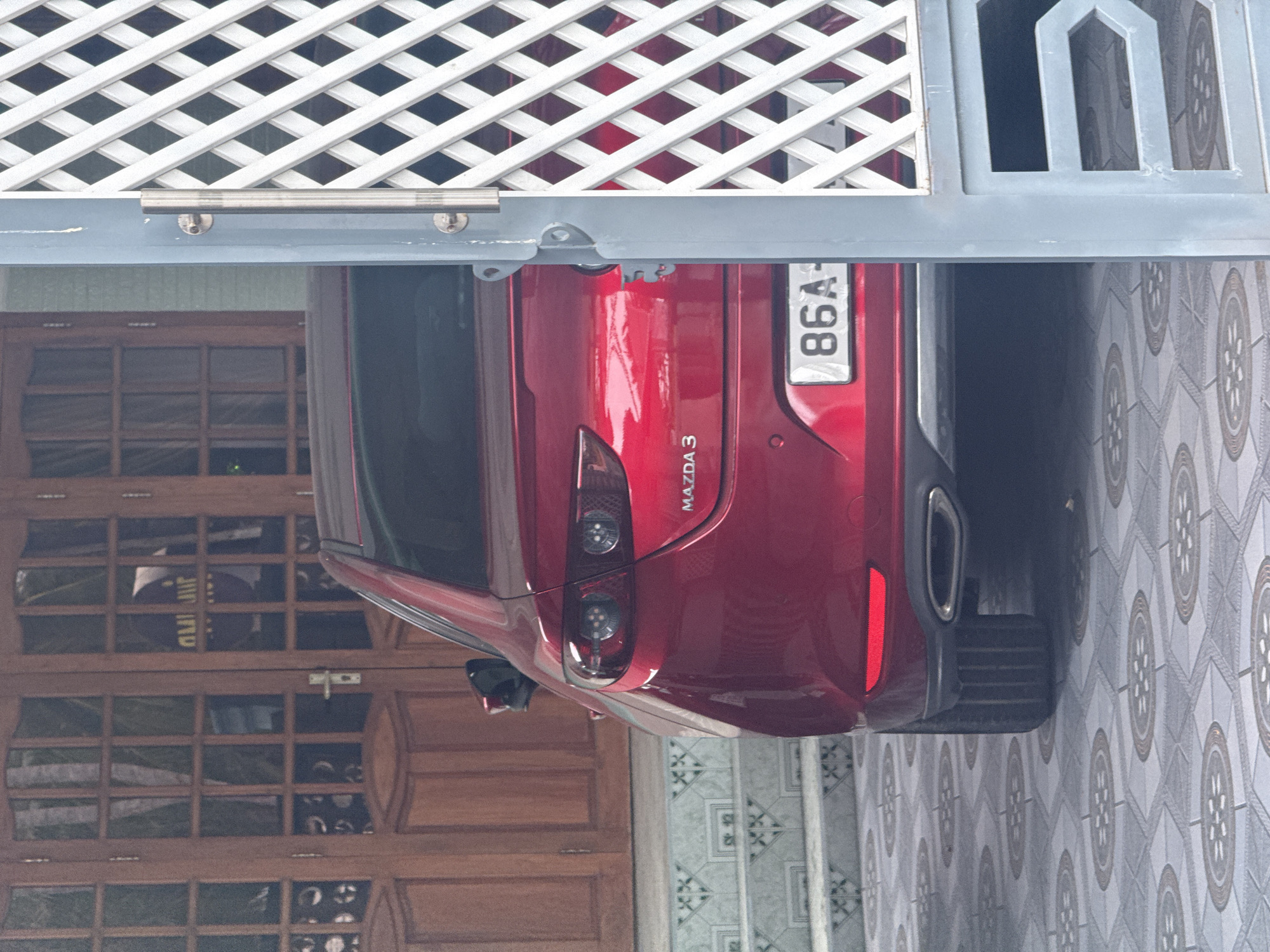 The image shows the rear of a red Mazda 3 parked in front of a wooden double door with glass panes. A light blue metal gate with a white lattice pattern is partially open on the right side of the frame, obscuring part of the car and the background. The car's license plate, partially visible, reads "86A-". The ground is tiled with a pattern of concentric circles. Steps with decorative tiles are visible to the left of the car. The scene appears to be in a residential or commercial area in Phu Thuy Ward, Vietnam. There are no people visible in the image.