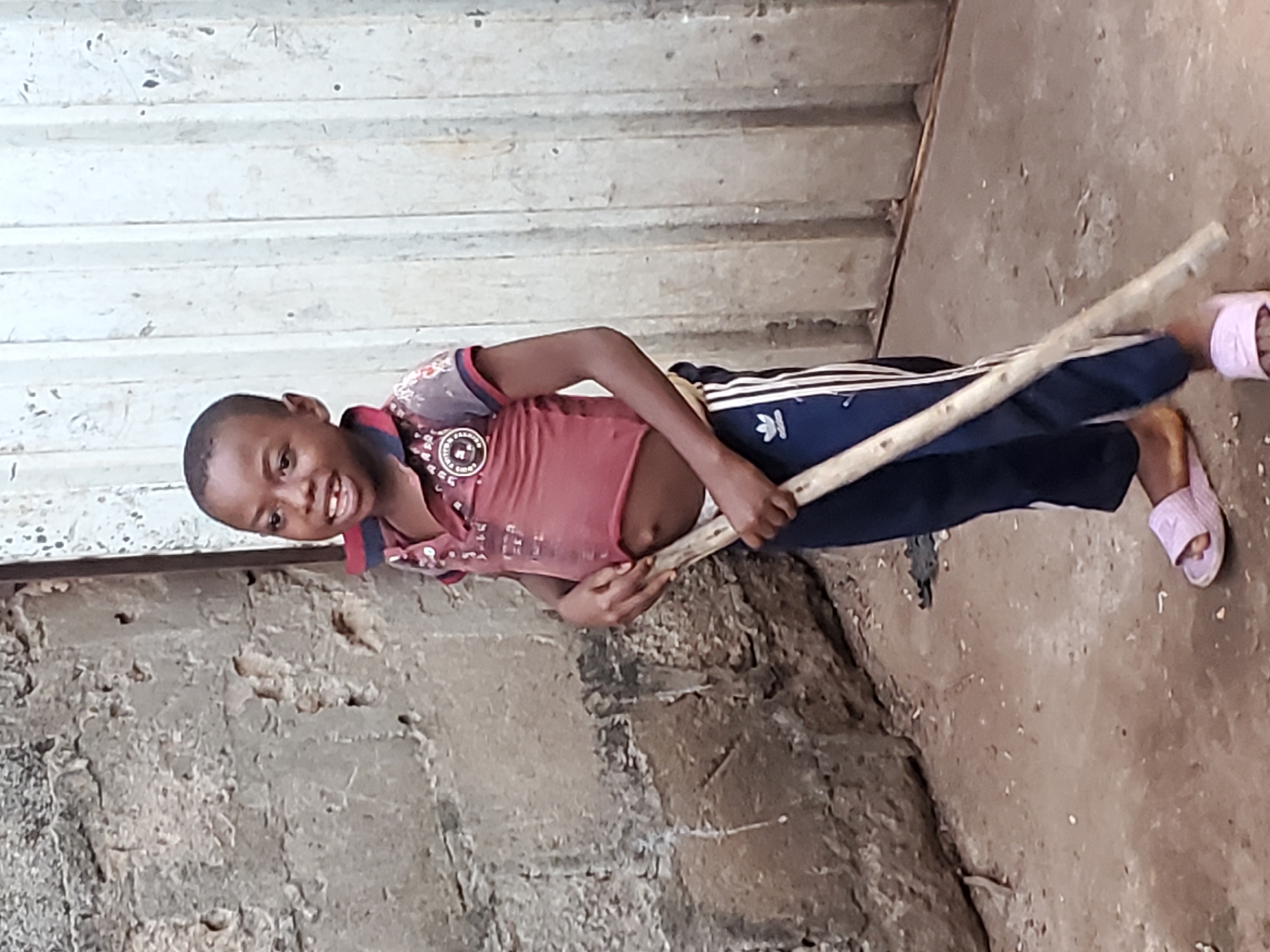 A young boy stands in front of a wall in Ukwu-Abwa, Nigeria. He is holding a wooden stick across his torso. He is wearing a red t-shirt, navy blue athletic pants with white stripes, and pink sandals. The wall behind him appears to be made of concrete blocks. The ground is dirt. A corrugated metal structure is in the background.