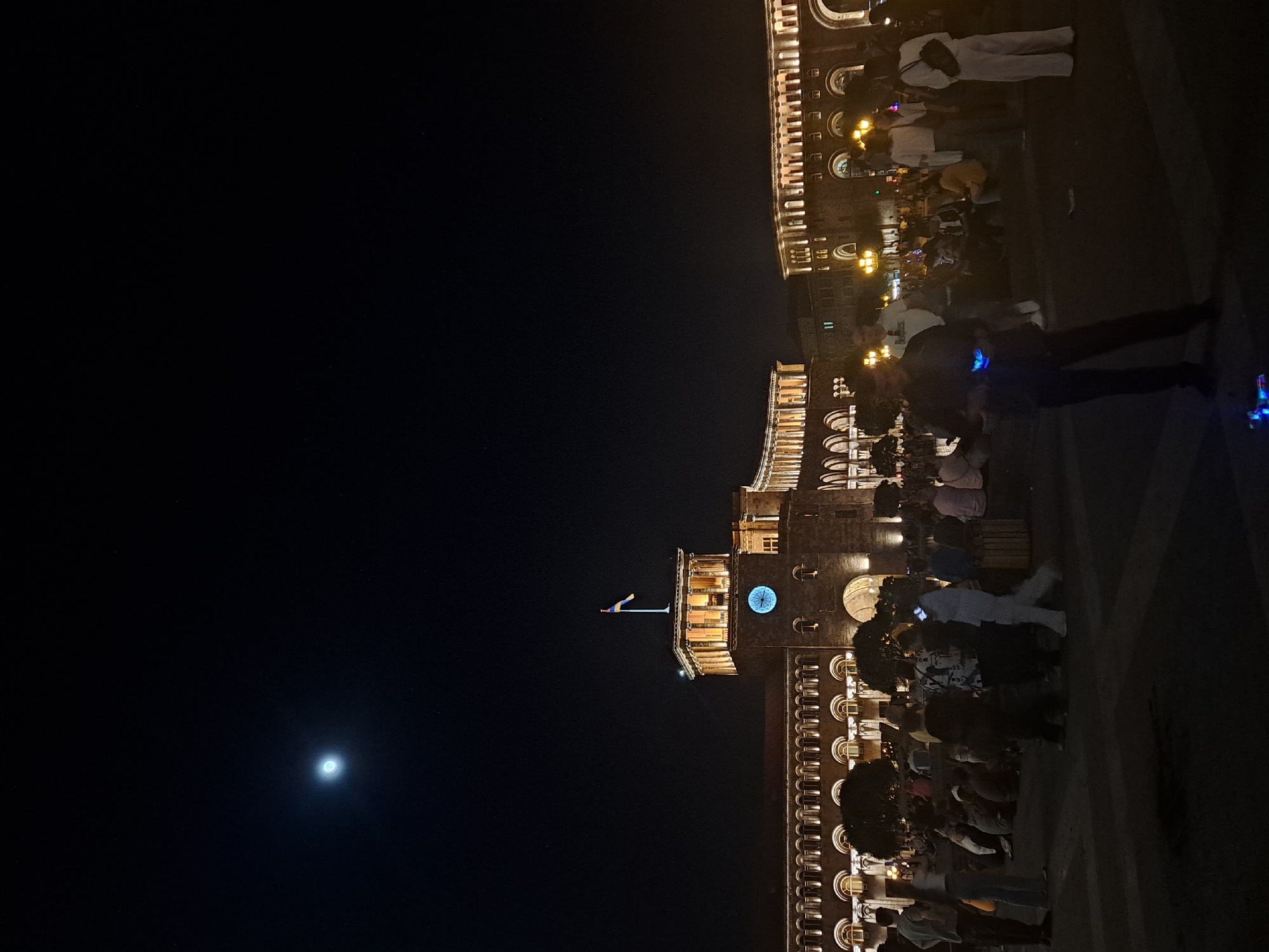 The image shows a night scene in Yerevan, Armenia, focusing on Republic Square. The sky is dark with a visible moon. Republic Square's main building is illuminated, featuring arched windows and a clock tower with a blue-lit clock face. The Armenian flag is visible atop the tower. The square is crowded with people, some standing and walking. The building's architecture and lighting are prominent.