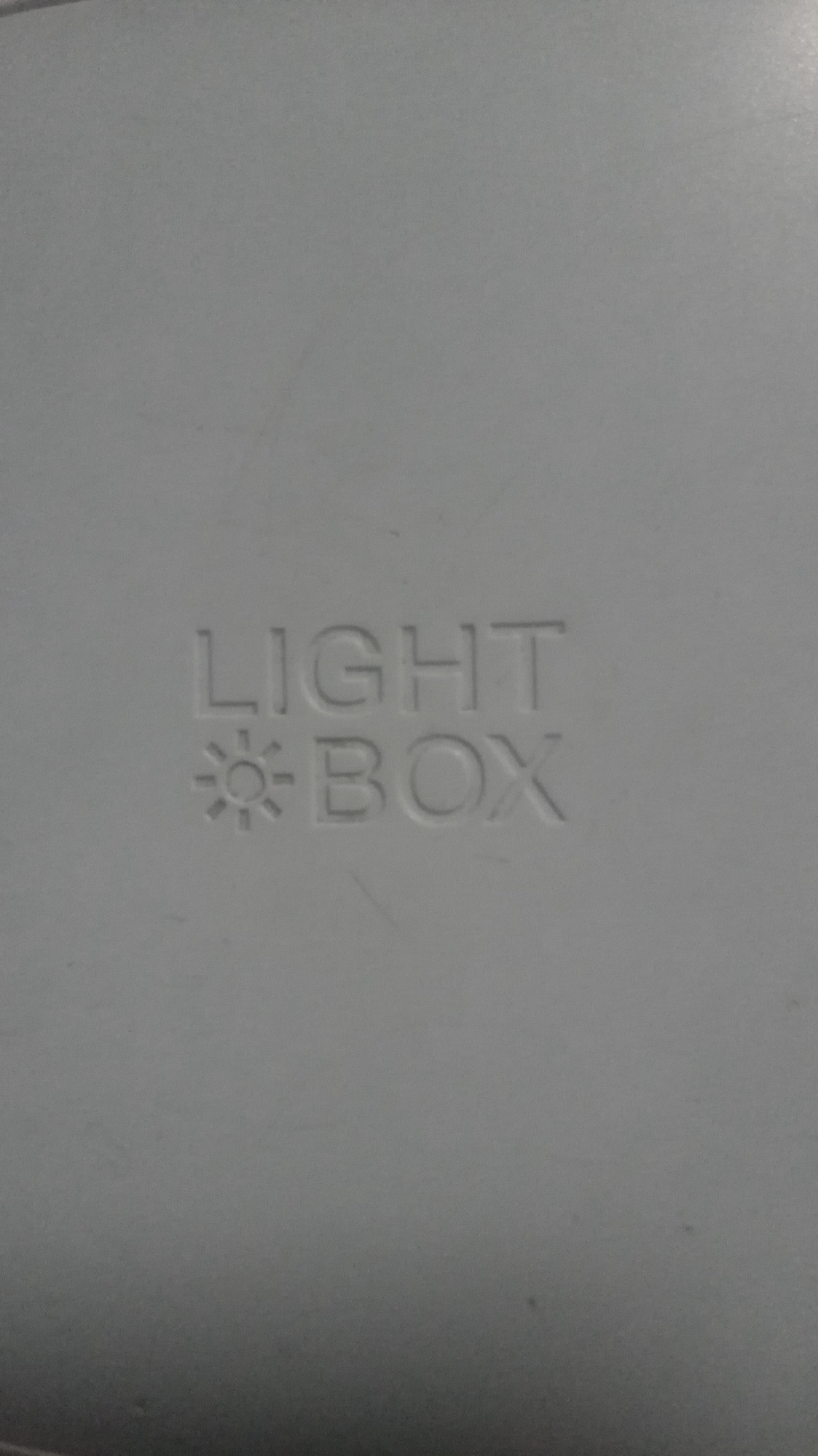 The image shows the text "LIGHT" above a symbol resembling a sun or light emitting object, which is above the word "BOX". The text and symbol are imprinted on a light gray or off-white surface. The lighting is uniform and the focal point is centered on the text.