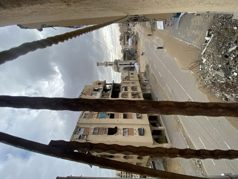 The image depicts a scene of devastation, viewed from an elevated perspective. Buildings are riddled with holes, and debris litters the street. A mosque's minaret stands amidst the destruction. The sky is overcast. Rust-colored metal bars and a rope occupy the foreground, framing the ruined cityscape. Two individuals can be seen walking on the street. The overall impression is one of conflict and urban ruin.