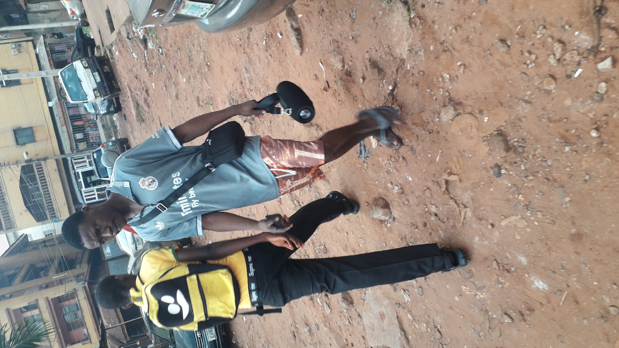 Two young men stand on a red dirt road in Onitsha, Nigeria. One wears a gray Real Madrid soccer jersey, orange shorts, and gray sandals. He carries a black messenger bag and a black speaker. The other man wears a yellow t-shirt, black pants, and black shoes. He carries a yellow backpack. Cars are parked on the side of the road, and buildings line the street.