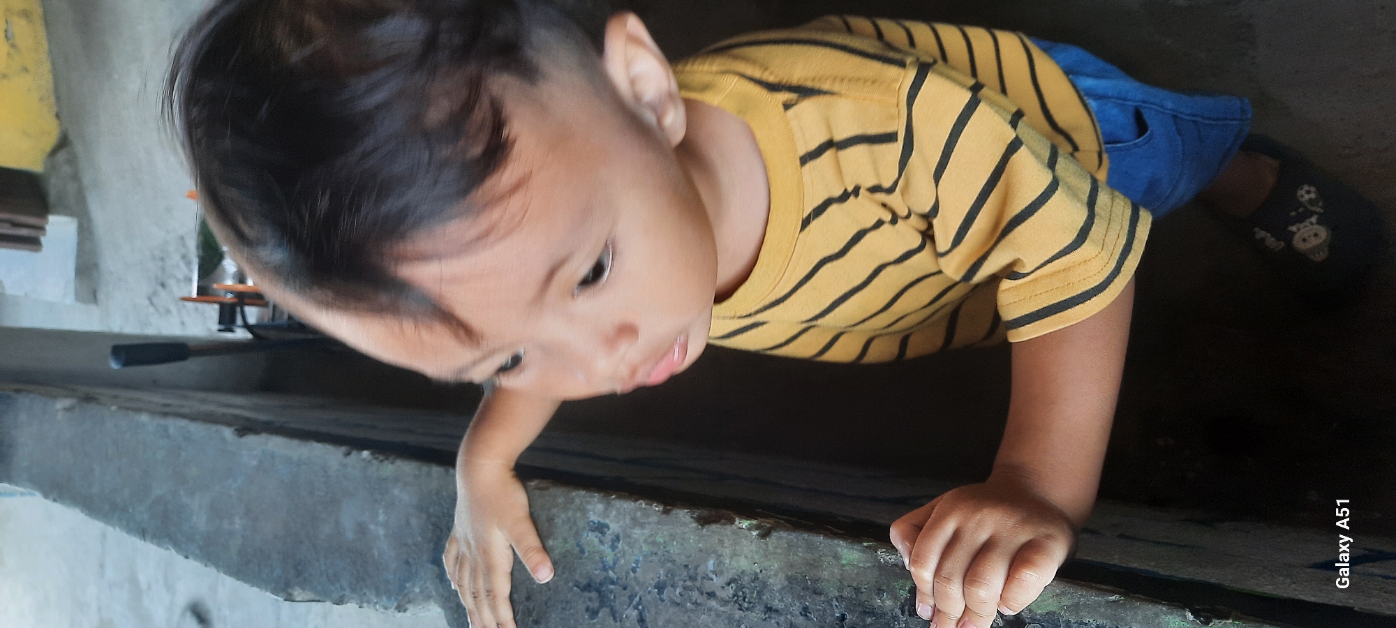 The image shows a close-up of a young child, likely a toddler, leaning over a rough, textured concrete ledge or wall. The child has dark hair, fair skin, and is looking downwards and to the left. They are wearing a yellow t-shirt with black horizontal stripes and blue jeans. The child's left hand is visible resting on the ledge, and their right arm is bent, also near the ledge. The background is dark and out of focus, with some indistinct shapes that suggest an outdoor or semi-enclosed area, possibly with a building. The presence of the "Galaxy A51" watermark in the lower left corner indicates the image was captured with a Samsung Galaxy A51 smartphone. This scene is located in Selopuro, Indonesia.