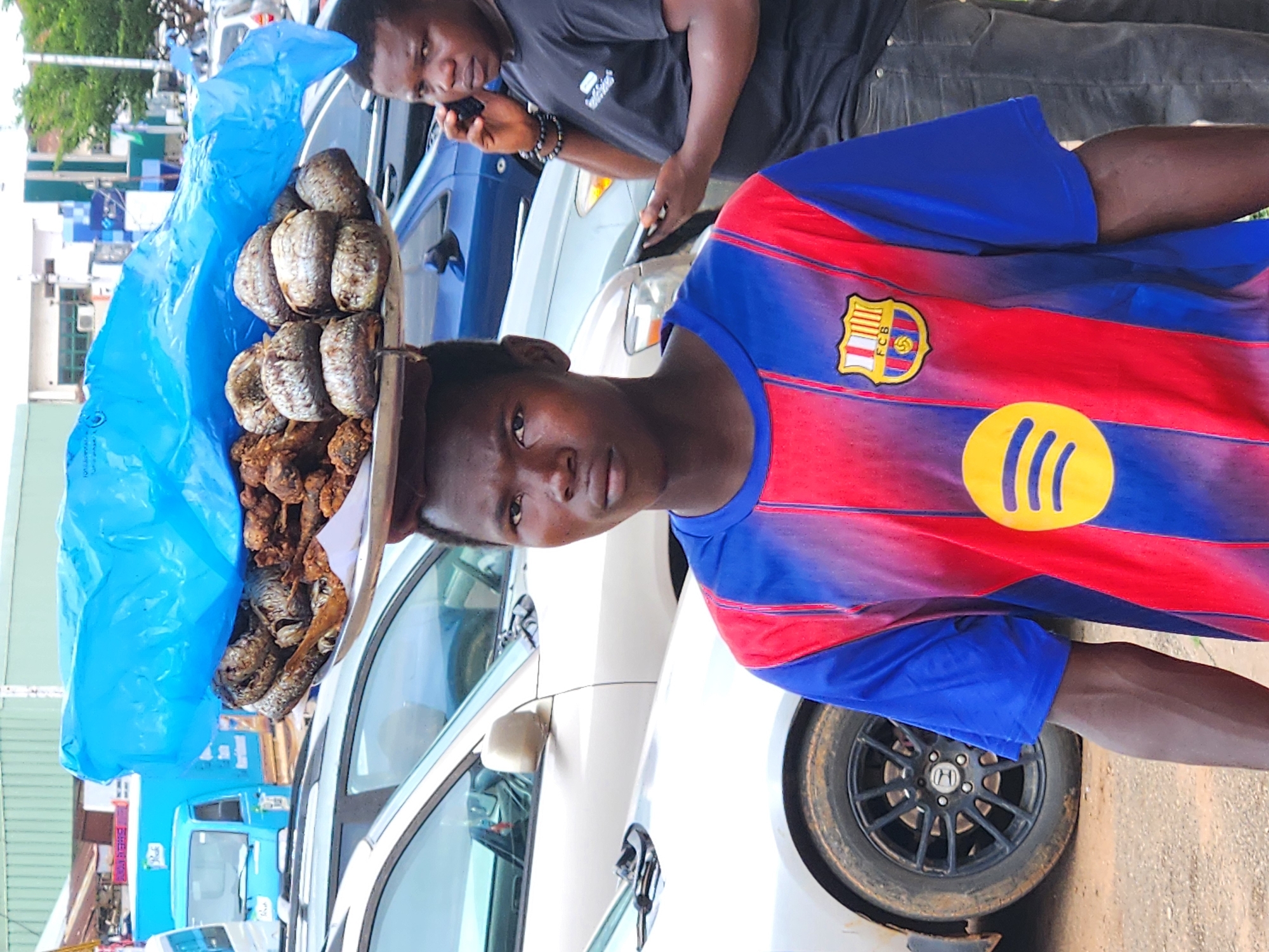 In an outdoor urban environment, a young man stands on a dirt road, carrying a tray of fried fish on his head, covered by a blue tarp. He is wearing a red and blue striped t-shirt with a crest and a yellow circular logo. In the background, a second man, wearing a black t-shirt, is talking on a mobile phone. Several cars, including a silver SUV and a blue sedan, are parked behind the young man. The scene appears to be a street market or roadside vendor area.