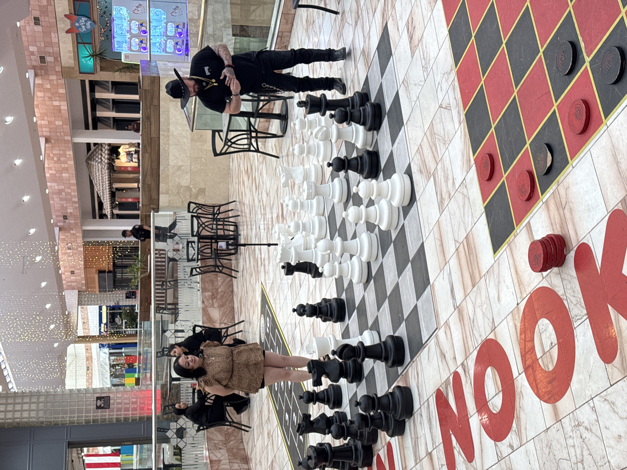 In El Paso, United States of America, within a well-lit indoor shopping mall, a large outdoor chess set is positioned on a checkered floor. The chessboard is approximately 30x30 feet, with alternating white and light brown marbled tiles. Several giant chess pieces, both black and white, are arranged on the board. In the foreground, to the right of the chessboard, a checkerboard pattern with red and black squares is visible, with red and black checkers placed on some of the squares.
In the midground, standing to the right of the chess set and facing it, is a man wearing a black cowboy hat, a black t-shirt, dark pants, and black boots. He has tattoos on his arms and is wearing a gold necklace. His hands are clasped in front of him. To the left of the chess set, a woman wearing a leopard-print dress and dark shorts is standing. Behind her, two other women in black attire are seated at a table.
In the background, the mall environment is visible with various shops, storefronts, and decorative lighting. Some tables and chairs for diners are also present. A security guard in a dark uniform is seen in the distance near a stairway. The scene suggests a recreational activity or a public gathering within the mall.