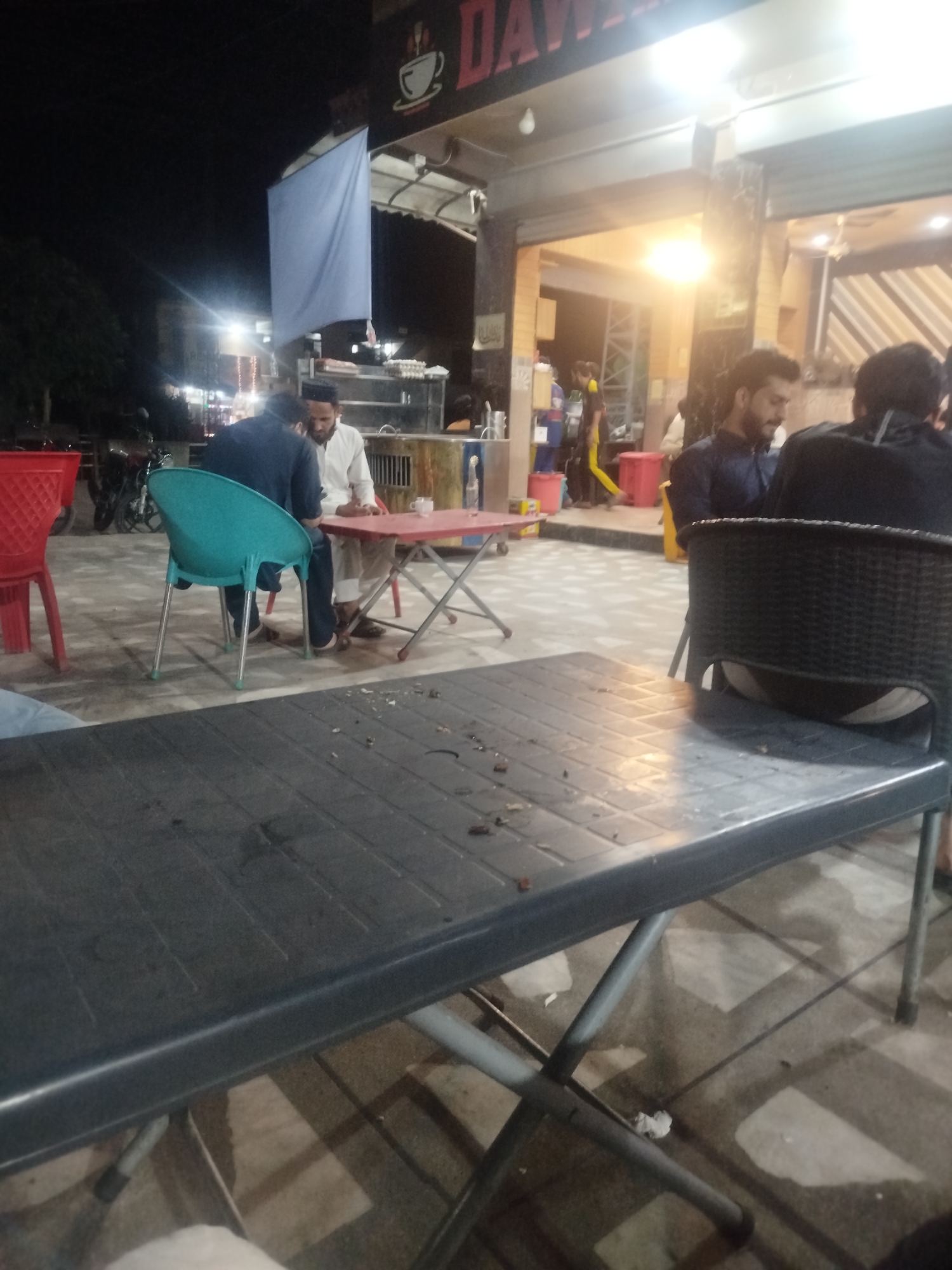 The image depicts an outdoor seating area at a restaurant or cafe in Peshawar City Tehsil, Pakistan. In the foreground, a dark gray plastic table with a metal folding frame is visible. Debris, possibly food scraps and cigarette butts, is scattered across the table's surface and the ground. In the mid-ground, several individuals are seated at tables. Two men in the center are facing each other at a red table, one wearing a white shalwar kameez and a skullcap, and the other in a dark blue shirt. A turquoise plastic chair is positioned in front of the man in the dark shirt. To the left, a red plastic chair is empty. Further back, other patrons are seated, and a food stall or kitchen area is partially visible. The setting appears to be at night, indicated by the artificial lighting and the dark sky. A large, pale blue sheet is hanging from an awning above the seating area. Motorcycles are parked in the background. The overall impression is a casual, open-air dining environment.