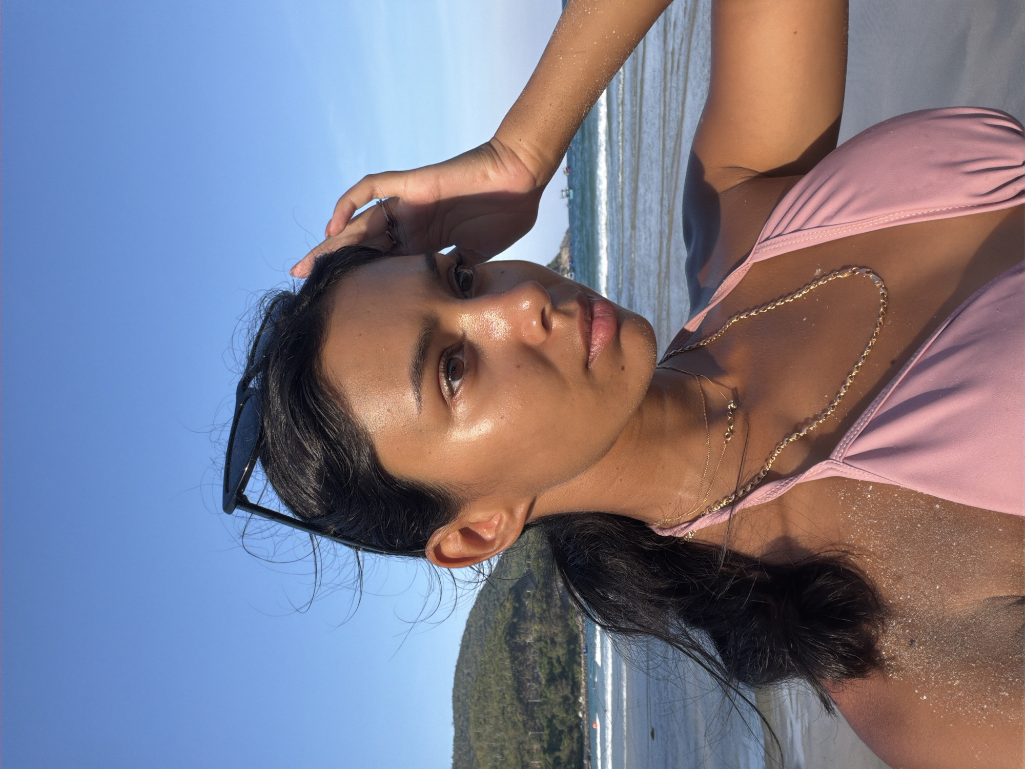 A medium shot depicts a woman at a beach in Nam Nha Trang Ward, Vietnam. She is wearing a pink swimsuit and gold necklaces. Sand is visible on her skin. Her dark hair is pulled back, with sunglasses tucked into it. She is touching her head with her right hand. The background shows the ocean, a beach, and a densely vegetated hill. The sky is clear and blue.
