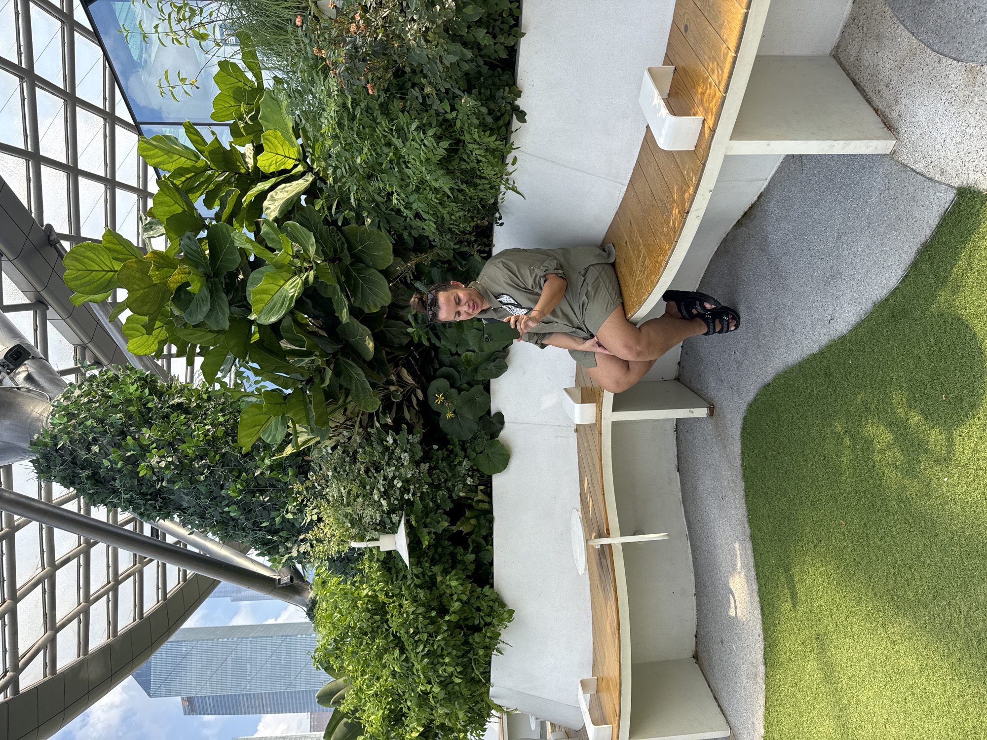 A woman sits on a curved bench constructed of white and wood materials, amidst lush greenery under a metal and glass ceiling, likely within an enclosed garden area. She is wearing an olive green short-sleeved shirt, shorts and black sandals. She is smiling, holding up two fingers, indicating a peace sign. The surrounding environment consists of various plants, including large-leafed trees. Artificial green grass is visible next to the bench. A view of the city skyline is seen through the glass ceiling, suggesting a high-rise building in Tianhe District, China.