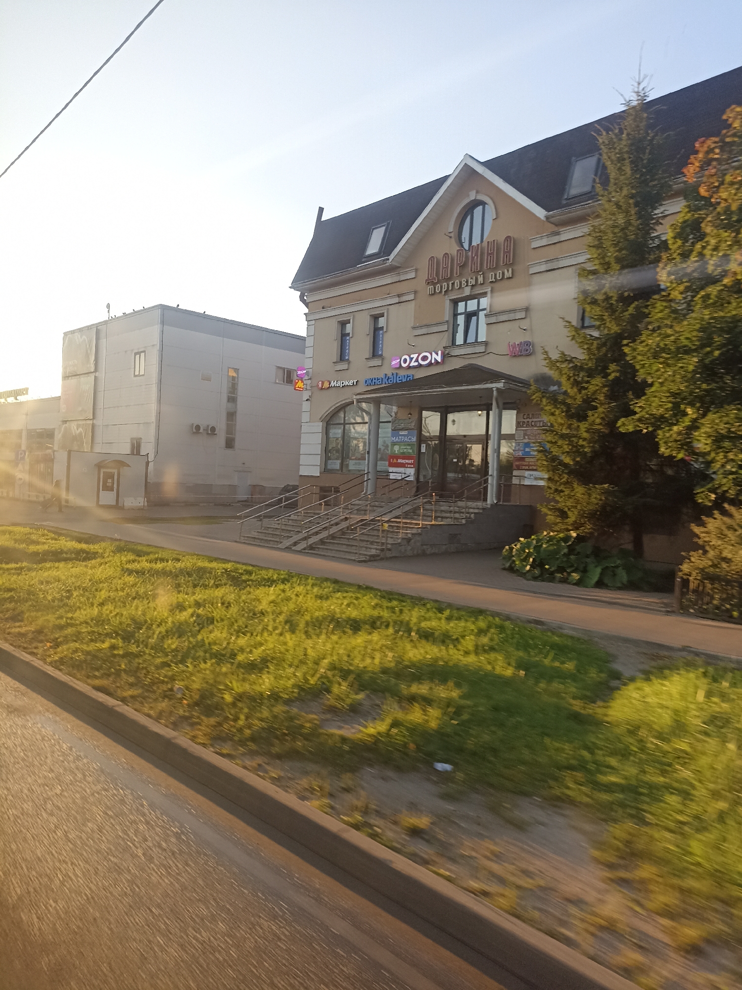 The image captures a sunny day in Chekhov, Russia, likely in the late afternoon due to the warm lighting. The scene features a commercial building with a light-colored facade and a dark roof, characteristic of many buildings in the region. Prominent signage on the building includes "ДАРИНА" (Darina) and "ТОРГОВЫЙ ДОМ" (Trading House) in Cyrillic, indicating a shopping center or marketplace. Below these main signs, smaller logos are visible, including "OZON," a popular Russian online marketplace, and "WB," likely referring to Wildberries, another e-commerce giant. The building has a glass-fronted entrance with stairs leading up to it, and several windows. Adjacent to this building is a taller, more modern structure, possibly an office or retail space, with a light gray exterior and several windows.
In the foreground, a patch of green grass, illuminated by the setting sun, borders a paved area, likely a sidewalk or road. A blurred car tire track is visible on the lower left, suggesting the photo was taken from a moving vehicle. The overall impression is one of a typical urban commercial district in a Russian city, bathed in the warm glow of natural light.
