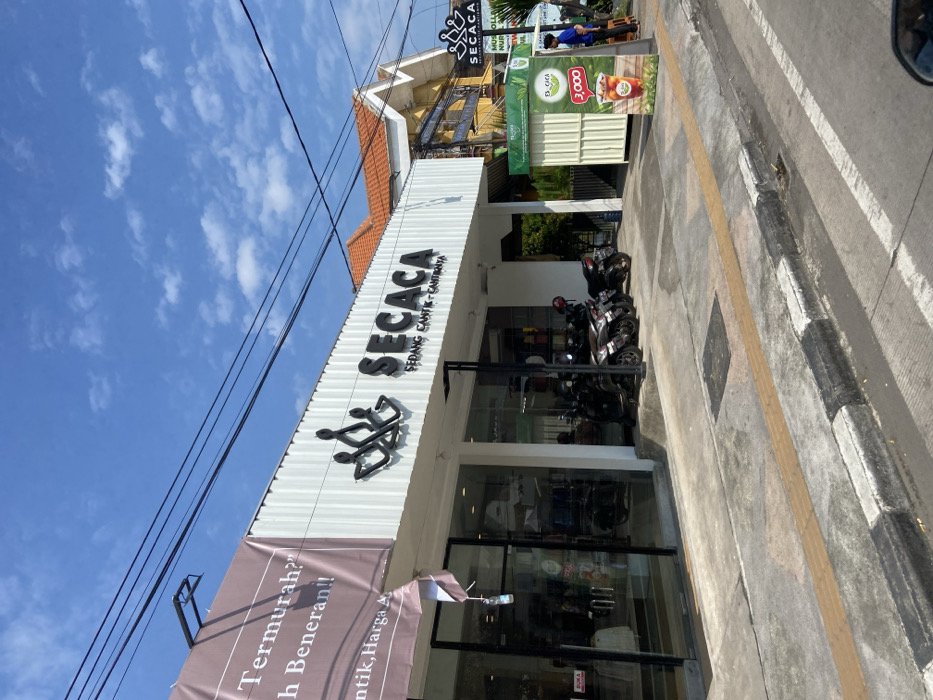 The image shows a storefront in Kecamatan Tulangan, Indonesia, at latitude -7.481 and longitude 112.645. The storefront is a white building with a sign that reads "SECACA SEDANG CANTIK CANTIKANYA". There is a smaller sign to the right of the storefront that also reads "SECACA". In front of the storefront, there are five parked motorbikes and a person standing in front of a small, white stall with a sign that reads "SECACA" and "3,000". The sidewalk and street are gray concrete. The sky is blue with white clouds. There is a pink sign in the image but it is partially obscured by the storefront. The pink sign has text in Indonesian and reads "Termurah? h Beneran!! ntik, Harga A".