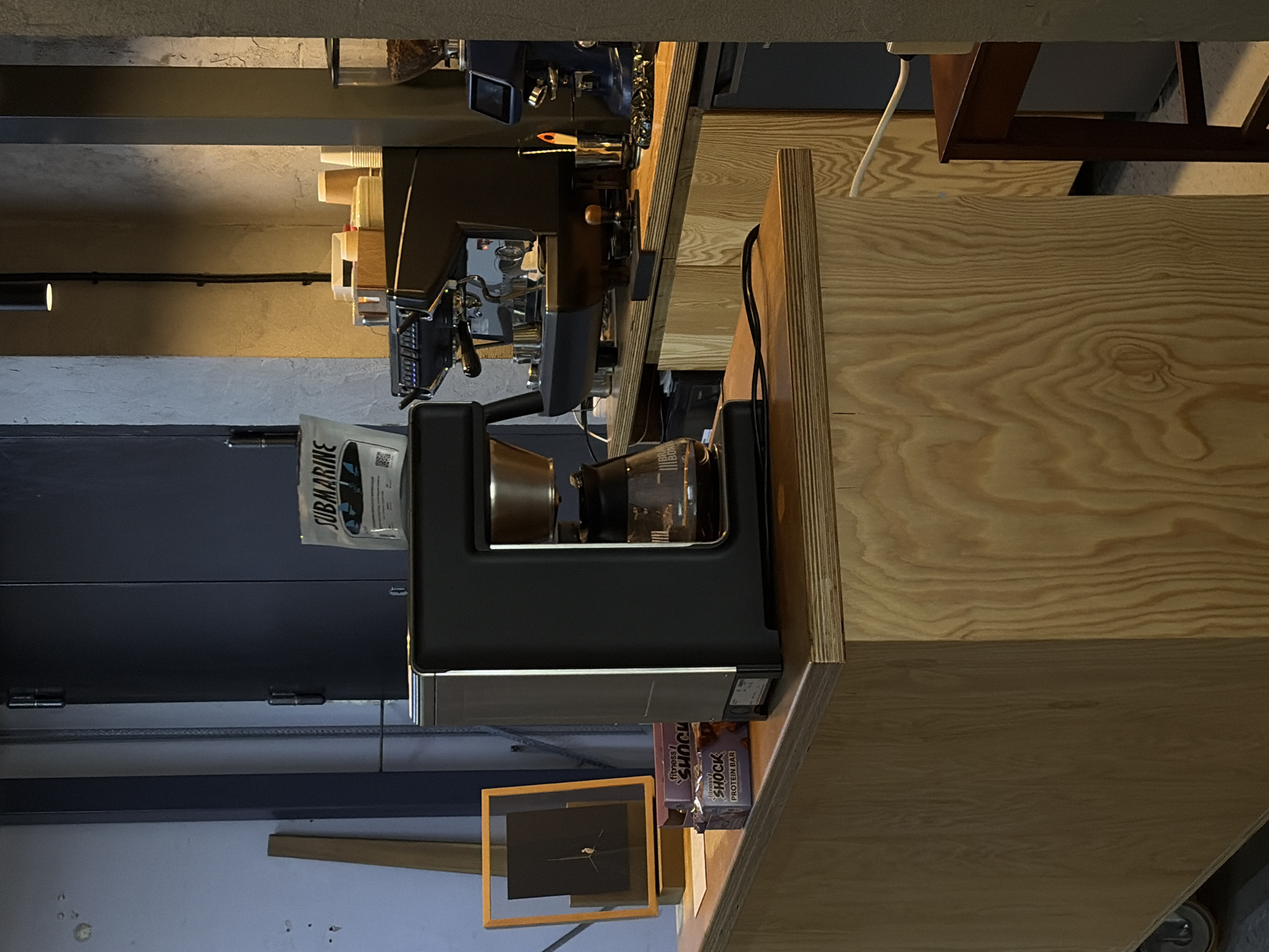 This is a medium shot, taken at eye level, of a coffee bar interior. The dominant feature is a light-colored wooden counter with a modern, black drip coffee maker on it. To the right of the coffee maker, there is a black espresso machine with a portafilter. Further to the right, a blue grinder is visible. On the counter, there are stacks of paper cups and other coffee-making paraphernalia. In front of the coffee maker, there are two purple packages of "Fitness Shock Protein Bar." A framed piece of art is to the left of the coffee maker. The background consists of a dark gray wall with exposed concrete texture and a large dark gray door. The lighting is dim, with a single pendant light visible in the upper right portion of the image. The image depicts a functional and stylish coffee bar, likely in a commercial establishment. No people are present in the image. The scene is clean and organized.
