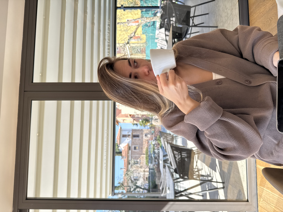 An image taken in Albisano, Italy, depicts a female subject seated indoors, consuming a beverage from a white ceramic mug. The subject has long, light brown hair with blonde highlights and wears a brown, ribbed cardigan over a white top. Her head is turned slightly to the left, and her gaze is directed away from the viewer. Her right hand holds the mug to her mouth.
The subject is positioned in front of a large window system, framed in dark brown, through which an outdoor patio and village scene are visible. The outdoor patio features multiple round tables with dark-colored, woven chairs. Beyond the patio, traditional Italian architecture is visible, consisting of multi-story buildings with terracotta roofs and light-colored facades, set against a clear blue sky. Sparse trees with some bare branches and some green foliage are also present. Reflected text, "AL CAMINETTO," is visible on the interior blinds or window structure above the outdoor scene. A portion of a dark-colored smartphone or electronic device is visible on a surface in the lower right foreground.