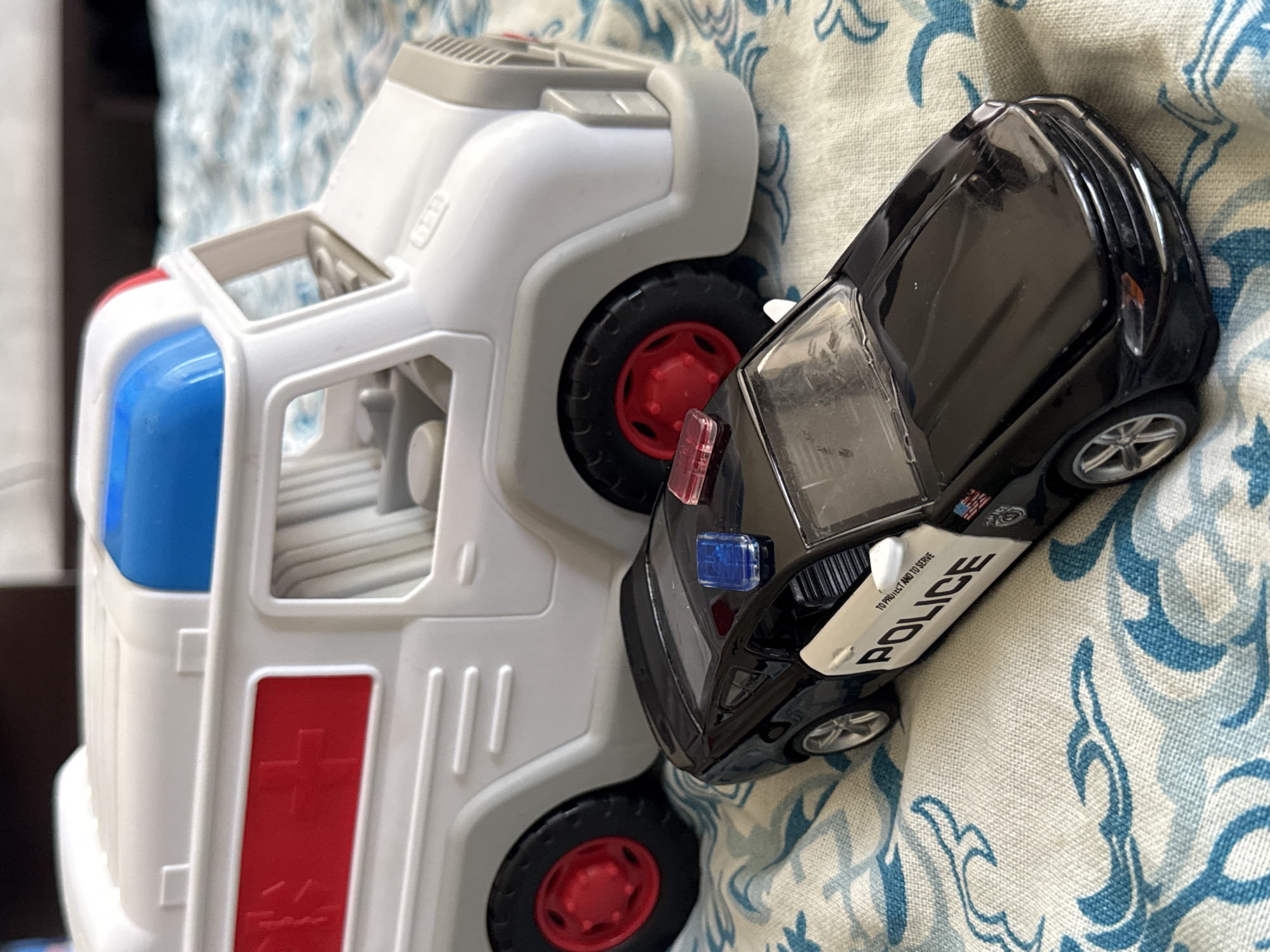 The image displays two toy vehicles, a police car and an ambulance, positioned on a patterned fabric surface. The police car, a black Ford Mustang replica, is in the foreground, facing towards the right. It features a white door with the word "POLICE" printed in black. The ambulance, a white toy truck with red and blue accents, is behind the police car, also facing right. The fabric surface has a blue and white swirling floral pattern. The scene appears to be indoors, likely a domestic setting. The location in Яромінскі сельскі Савет, Belarus is not discernible from the image itself.