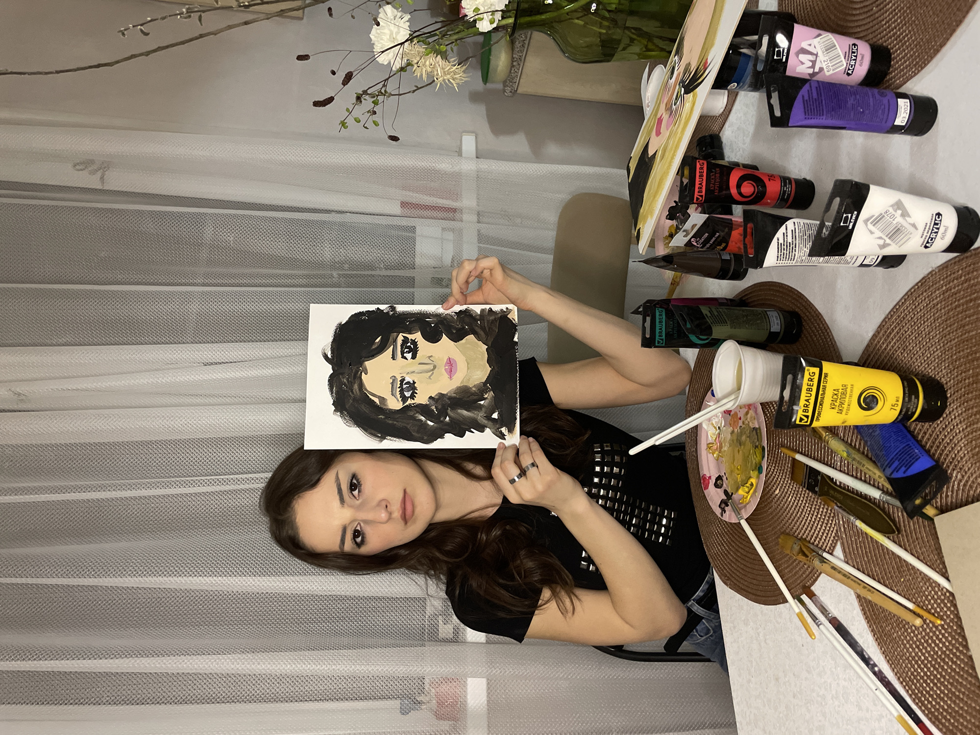A young woman with long, dark, wavy hair is pictured indoors, seated at a table in Khimki, Russia. She is holding up a small portrait painting for the camera, showcasing her artwork. The painting depicts a stylized face with dark, curly hair, light skin, prominent eyes with dark lashes, and pink lips, rendered in an expressive style. The woman looks directly at the viewer with a neutral expression.
Surrounding her on the table are various art supplies, including multiple tubes of acrylic paint, several paintbrushes, a small palette containing yellow and pink paint mixtures, and a cup of water. These items rest on a round, woven placemat. In the background, sheer white curtains are visible, along with a vase holding white flowers and some bare branches. The indoor lighting suggests a potentially evening or night setting. The woman is wearing a dark top adorned with metallic studs and a ring on her left hand.
Visible text on the paint tubes includes brands "BRAUBERG" and "MALEV". Descriptions such as "ПРОФЕССИОНАЛЬНАЯ СЕРИЯ КРАСКА АКРИЛОВАЯ ХУДОЖЕСТВЕННАЯ" (Professional series acrylic artist's paint) and "Матовая" (Matte) are present, along with volumes "75 мл" and "60ml", product codes such as "1078" and "6917", and a date "03.2021".