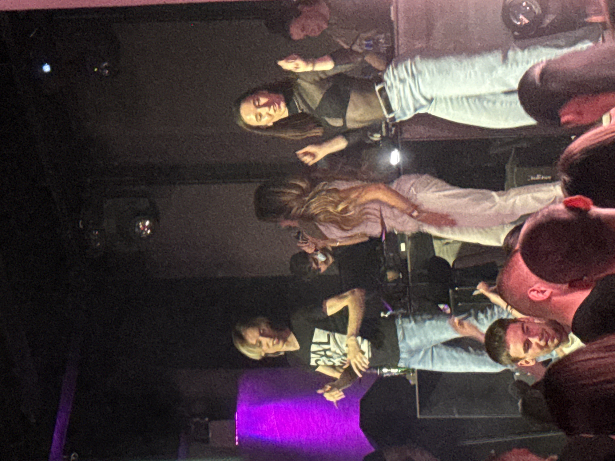 The image shows a live music performance indoors, possibly in Varna, Bulgaria. A band or group of performers is on stage, lit by purple stage lighting. A male singer is front and center, wearing a black T-shirt with white writing and light-colored jeans. To the left of the image, a female performer with long dark hair is visible, wearing a black top and light jeans. Another person is behind them and towards the center with longer lighter colored hair, wearing a light pink long-sleeved top and white pants. The scene is crowded, with audience members visible in the foreground, mainly heads and shoulders. The background is dark, suggesting a dimly lit club or venue. Audio equipment, including speakers and amplifiers, can be seen behind the performers on stage.