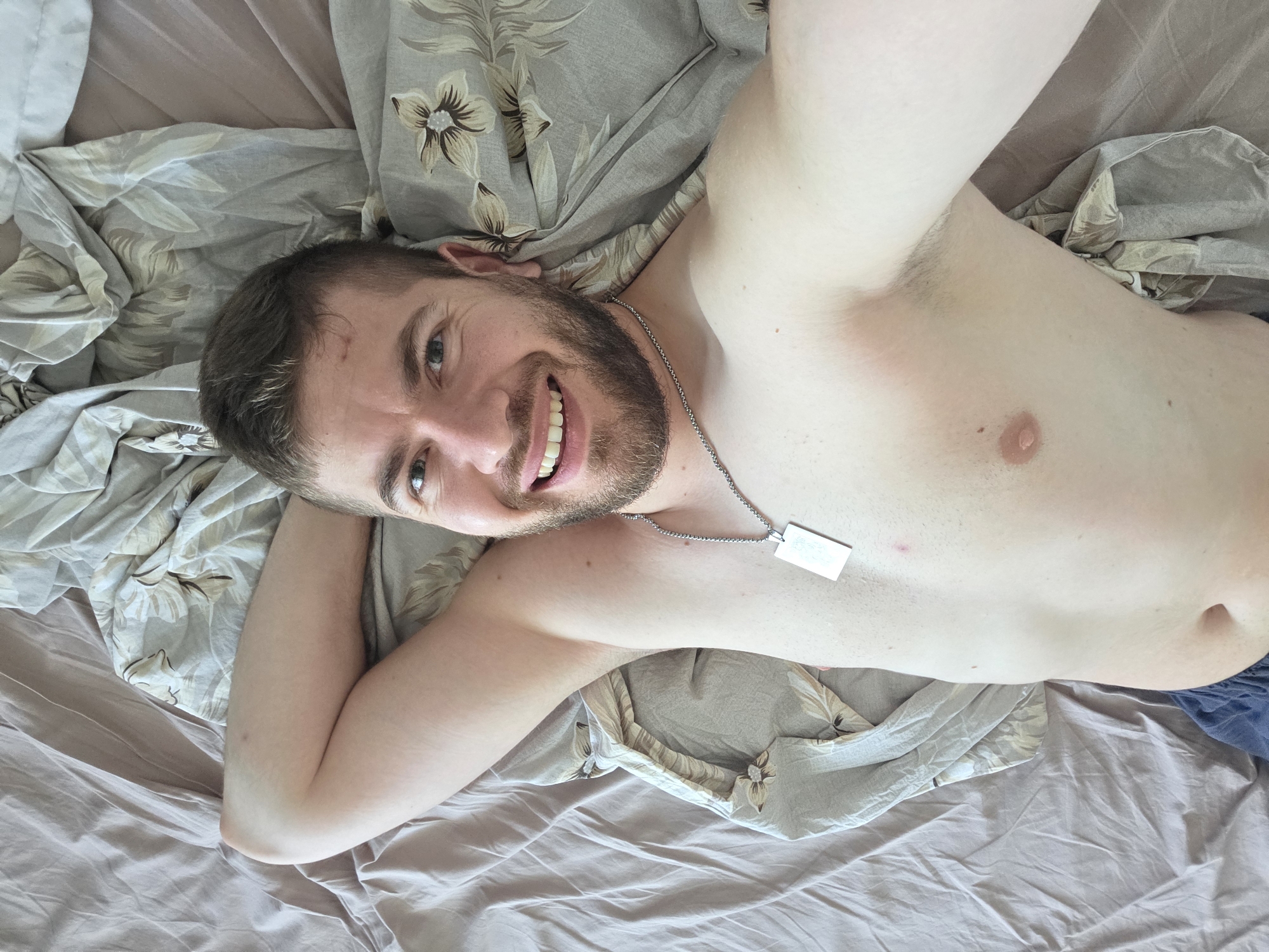 A fair-skinned man with short brown hair and a beard lies on a bed with patterned and solid-colored linens. He is shirtless and wears a silver chain with a rectangular pendant. The location is not discernible from the image. No actions or interactions are visible. The environment consists of the bedding.