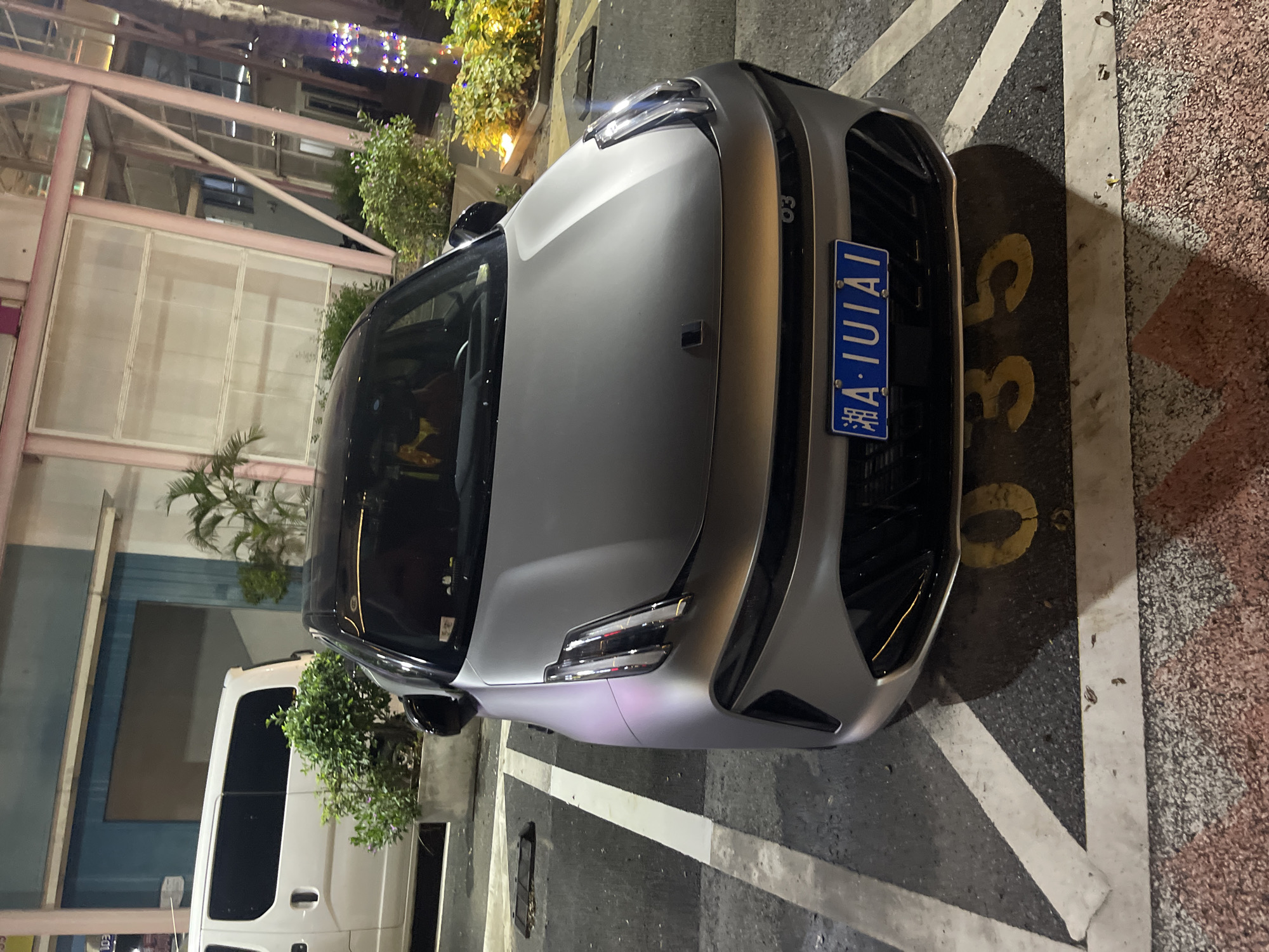 The image captures a silver car parked in a lot at night. The car is positioned facing forward, with its front bumper and headlights prominently displayed. A blue license plate with white lettering is attached to the front of the car, reading "A-1UIAL". The parking space is marked with white lines, and the number "35" is visible in yellow paint on the ground next to the car.
In the background, a building with a pink metal structure and glass panels is visible, illuminated by artificial lights. There are also lush green plants and trees surrounding the building and the parking lot. To the left of the silver car, a white van is partially visible, also parked. The overall scene suggests an urban or commercial setting in Panyu District, China, during nighttime. The lighting suggests it is evening or night. There are no people visible in the image.