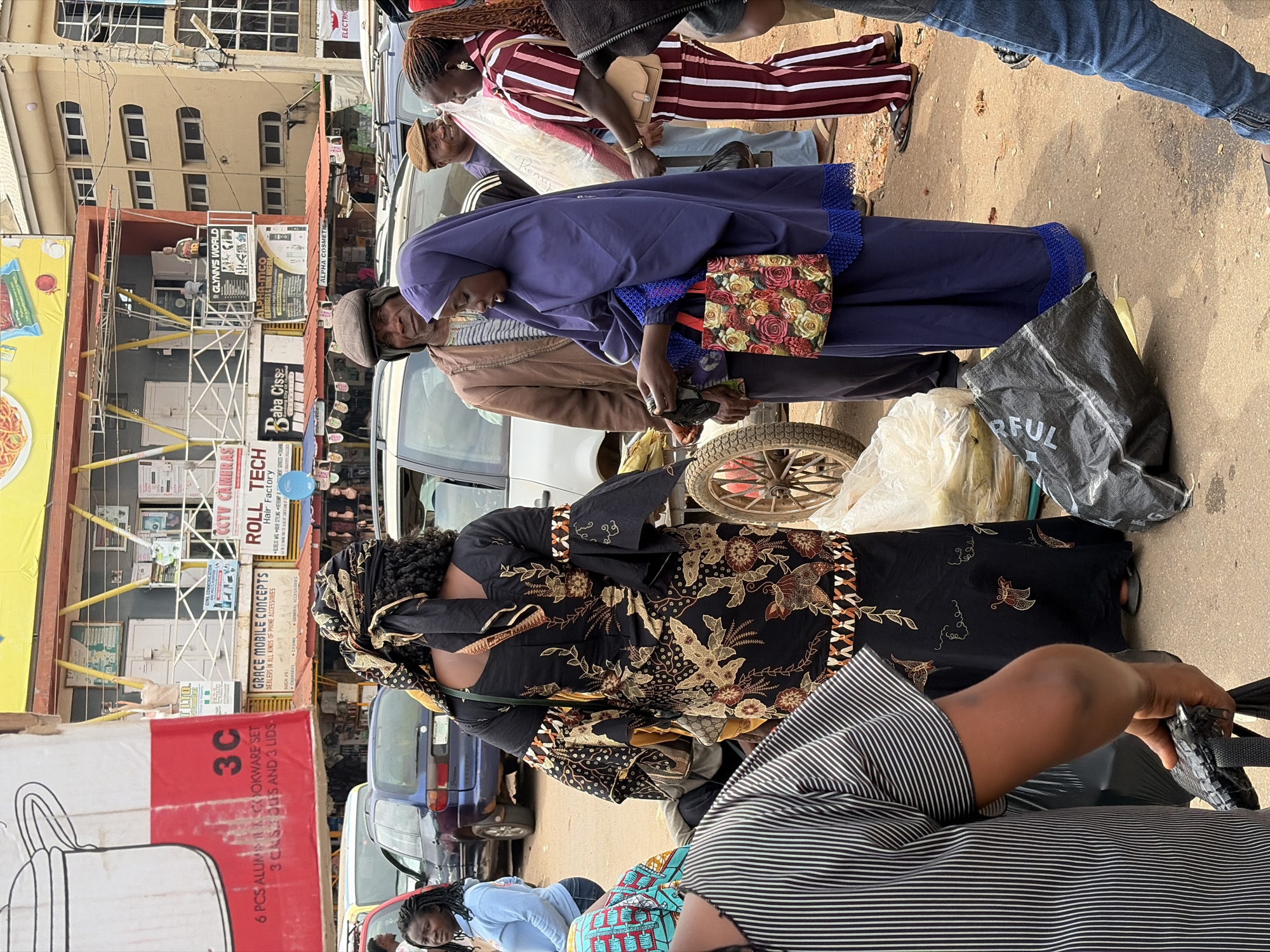 This is a street scene in Jos, Nigeria, likely a market area, with a variety of people and vehicles. The foreground shows a woman in a black dress with a gold and red floral pattern and a headscarf, standing next to another woman in a purple hijab and a floral patterned tote bag. A man in a brown jacket and flat cap is also present. To the left, a person wearing a black and white striped shirt is partially visible, holding a black bag. In the background, there are several cars, including a blue SUV and a silver minivan. Above the street level, buildings with signs are visible, including "ROLL TECH Hair Factory" and "CCTV CAMERAS." The ground is paved and dusty. The lighting suggests it is daytime.