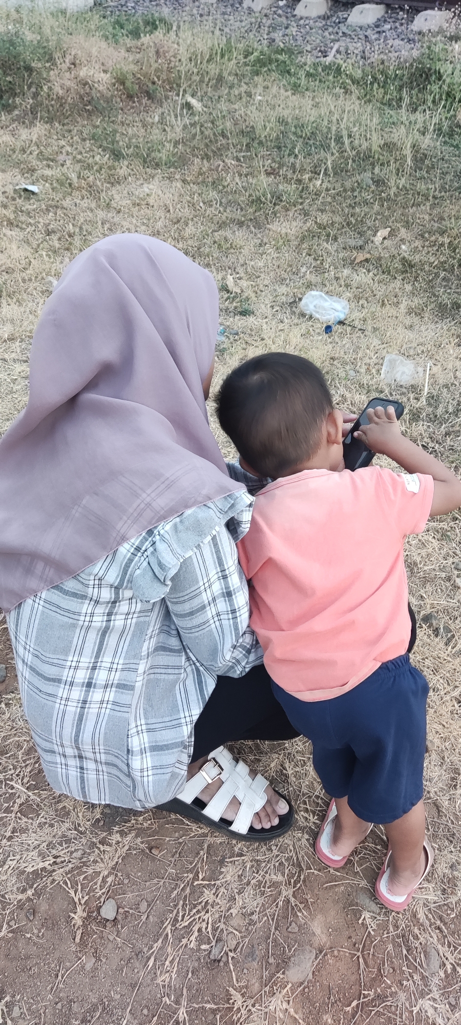 Two individuals, an adult and a child, are seated on dry, light-colored grass and dirt. The adult, viewed from behind, wears a lavender hijab and a gray and white plaid long-sleeved shirt. White sandals are on their feet. The child, also viewed from behind, wears a peach-colored t-shirt and dark blue shorts. They are holding a black mobile phone to their ear. The background is predominantly dry grass and sparse vegetation. In the upper portion of the image, a few concrete railroad ties and rusted metal tracks are visible, suggesting the scene is located near a railway line. The general environment appears rural or semi-rural, consistent with the outskirts of Pakusamben, Indonesia.