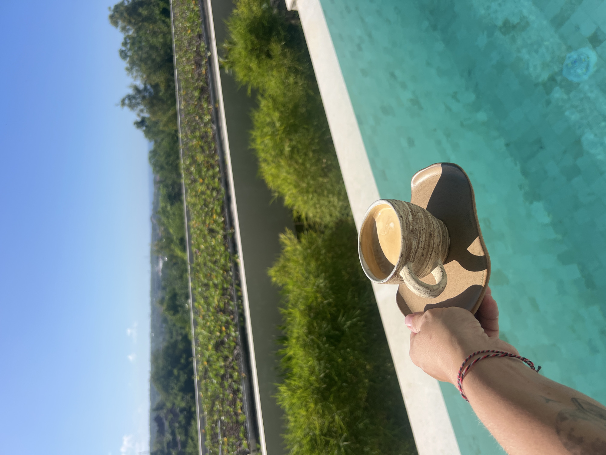 A first-person perspective image depicts a hand holding a mug of coffee on a saucer next to a light blue pool, likely in Kuta Selatan, Indonesia, given the tropical vegetation and architectural style. The hand wears a red and black braided bracelet and has a tattoo. In the background, there are green plants growing on a vertical wall, separated by white dividers. A blue sky is visible in the upper left portion of the frame.