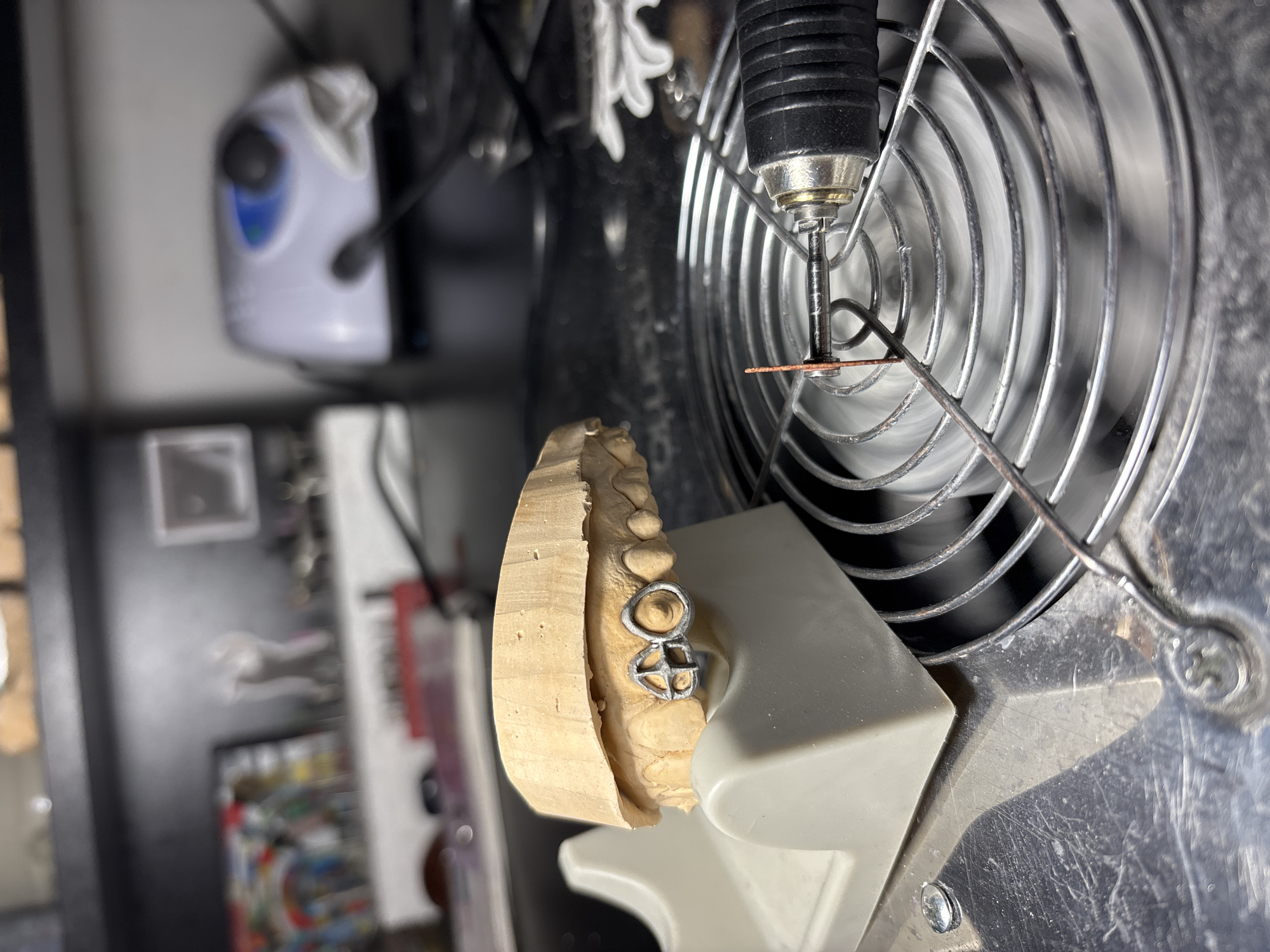 The image shows a dental model with a silver dental appliance attached, positioned in front of a rotary tool with a small grinding wheel. The rotary tool appears to be shielded by a metal fan guard. The scene is cluttered with other dental or technical equipment, but they are out of focus. The location is unknown.