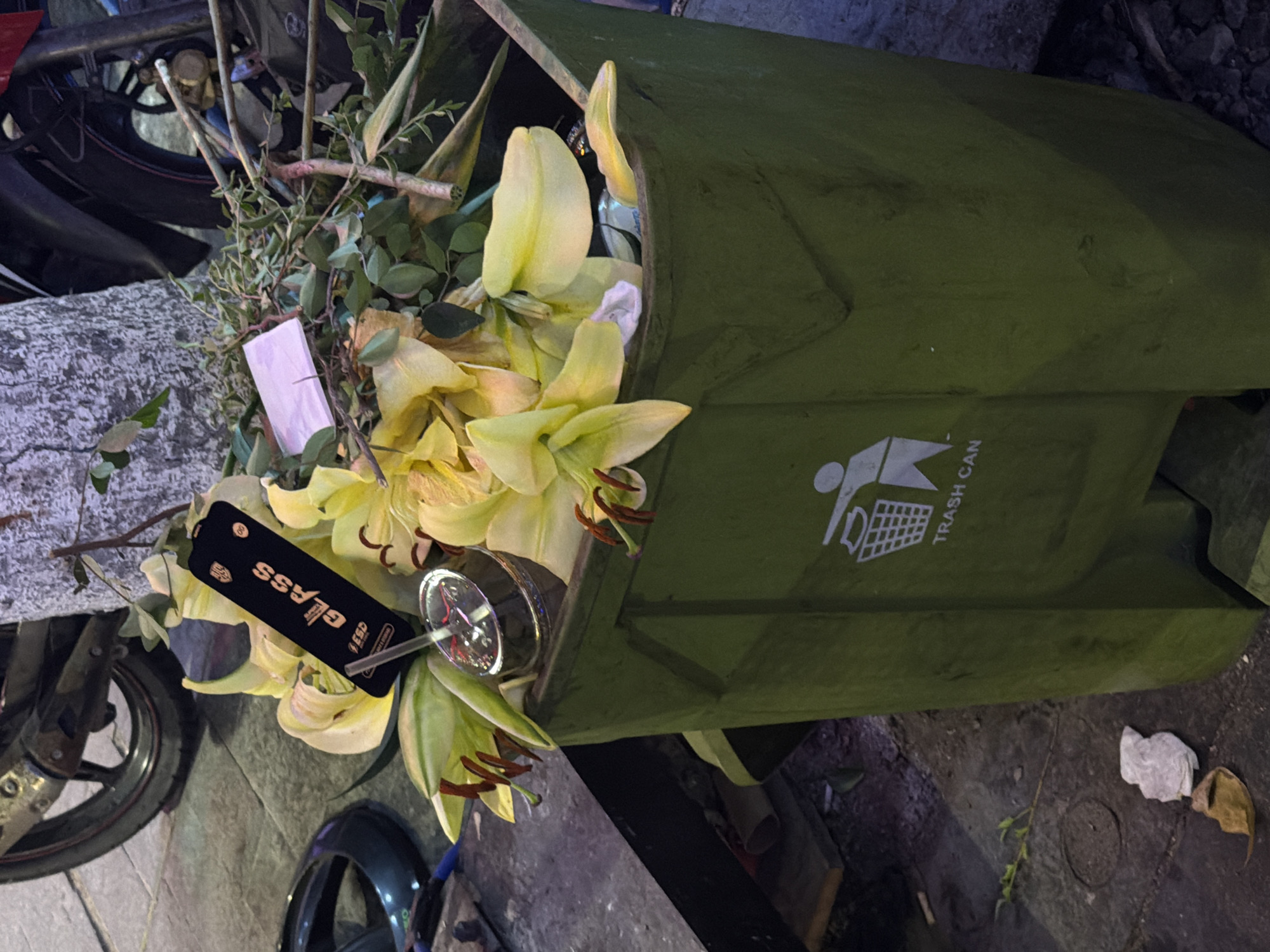 A green trash can overflowing with yellow lilies, branches, and a wine glass sits beside a motorcycle on a stone surface in Đà Nẵng, Vietnam. A "GLASS" tag is partially visible near the wine glass. Litter is scattered on the ground.