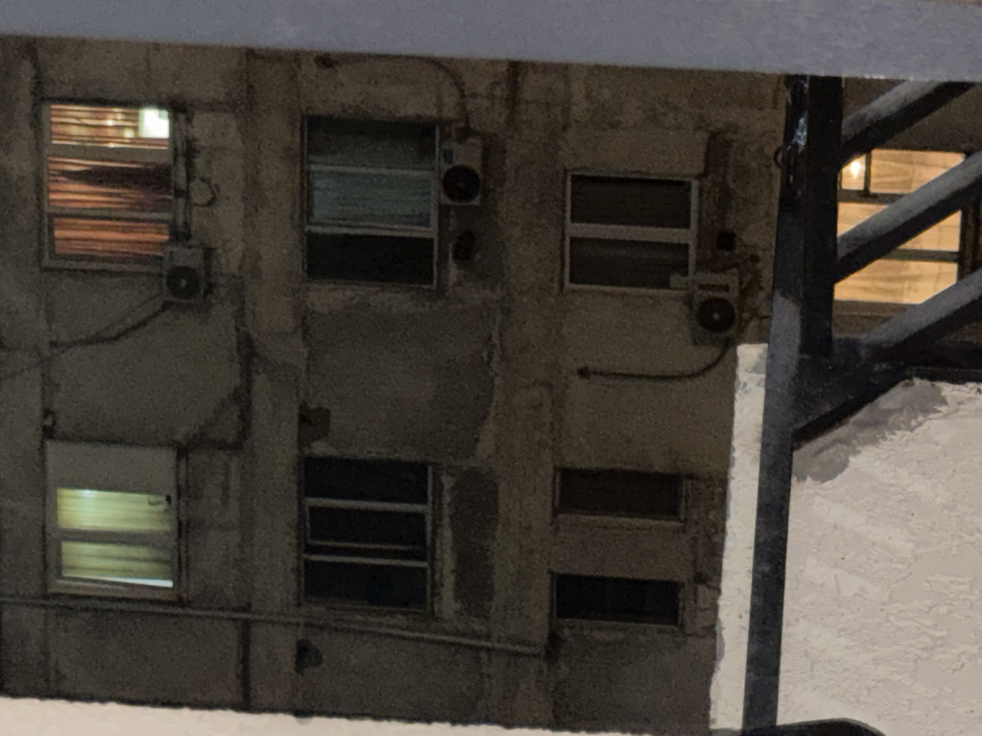 The image shows a close-up view of a residential building at night. The building appears to be made of concrete with a rough texture, and several windows are visible. Some windows have curtains, and lights are on in some of the rooms, casting a warm glow. There are also air conditioning units mounted on the exterior walls of the building. In the foreground, a dark metal railing is visible, suggesting the vantage point is from a balcony or similar structure. The overall scene depicts a quiet, urban nighttime environment.