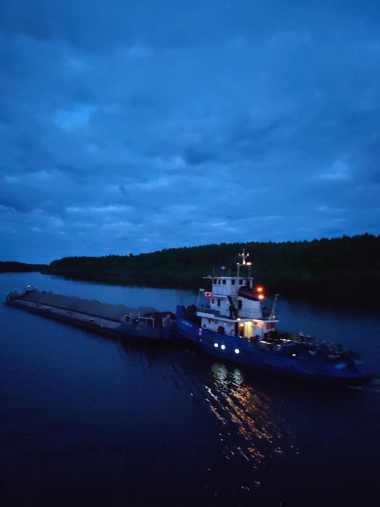 The image depicts a barge being towed by a tugboat on a waterway, likely a river or canal, in Alexandrovskoye, Russia. The tugboat is brightly lit with multiple lights, reflecting on the water surface. The barge is loaded with a dark, granular material, possibly gravel or sand. The scene appears to be at dusk or night, with a dark, cloudy sky overhead and wooded banks lining the waterway.