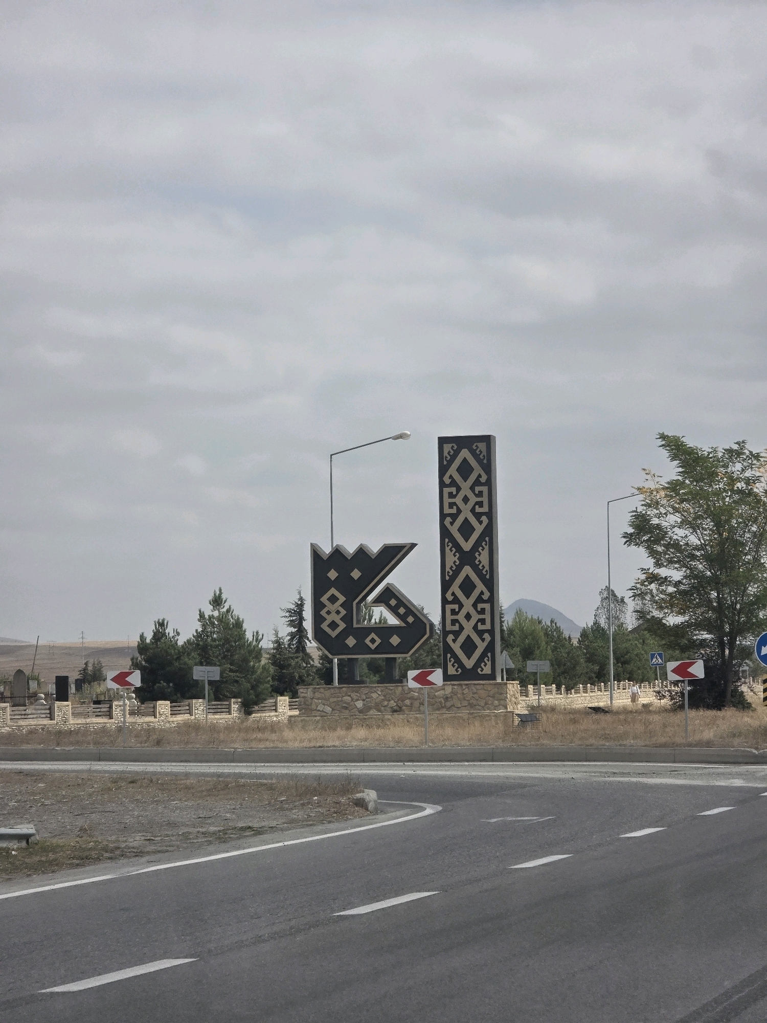 The image captures a large, abstract sculpture situated near a road intersection in Sabir, Azerbaijan. The sculpture is composed of two main elements: a stylized, crown-like shape with a black and patterned design, and a tall, rectangular pillar also adorned with a black and patterned motif. The sculpture is placed on a stone base and is surrounded by a low, decorative stone wall.
To the left of the sculpture, there are several evergreen trees and a sign with a red and white arrow pointing left. Further left, there appears to be a cemetery with several tombstones and a fence. A street light stands behind the sculpture.
To the right of the sculpture, there is a large deciduous tree with green foliage. Another street light is visible behind it. Further to the right, there are road signs, including one with a blue circle and a white arrow indicating a directional path, and another with a red and white arrow pointing right. A person is walking in the distance on the right side of the image.
The environment appears to be arid, with dry grass and sparse vegetation in the foreground. The ground is covered with asphalt for the road, marked with white lines. In the background, there are rolling hills and mountains under a cloudy sky. Power lines are visible in the far distance on the left. The overall scene suggests a roadside monument or landmark in a somewhat rural or semi-urban setting.