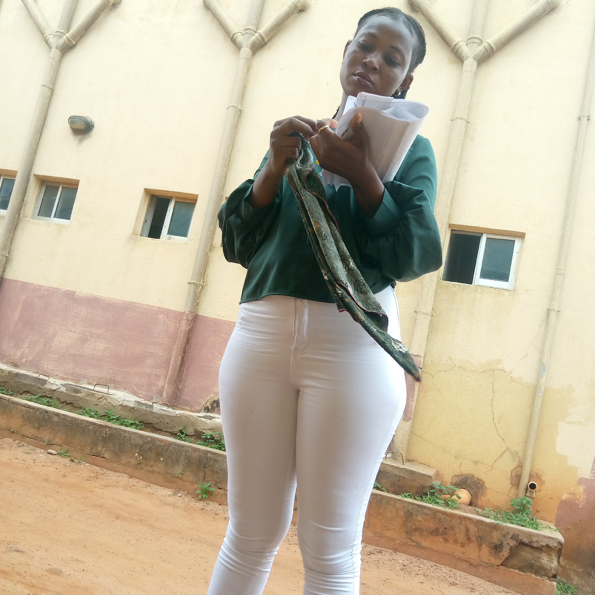 A woman stands on a dirt surface adjacent to a building with a cream-colored exterior, brown accents, and several windows. She is wearing a dark green top with long, puffy sleeves and white pants. She holds papers in her right hand and a patterned scarf in her left. The location is Ukwu-Abwa, Nigeria. The setting appears to be an outdoor space next to a building with water pipes and windows.