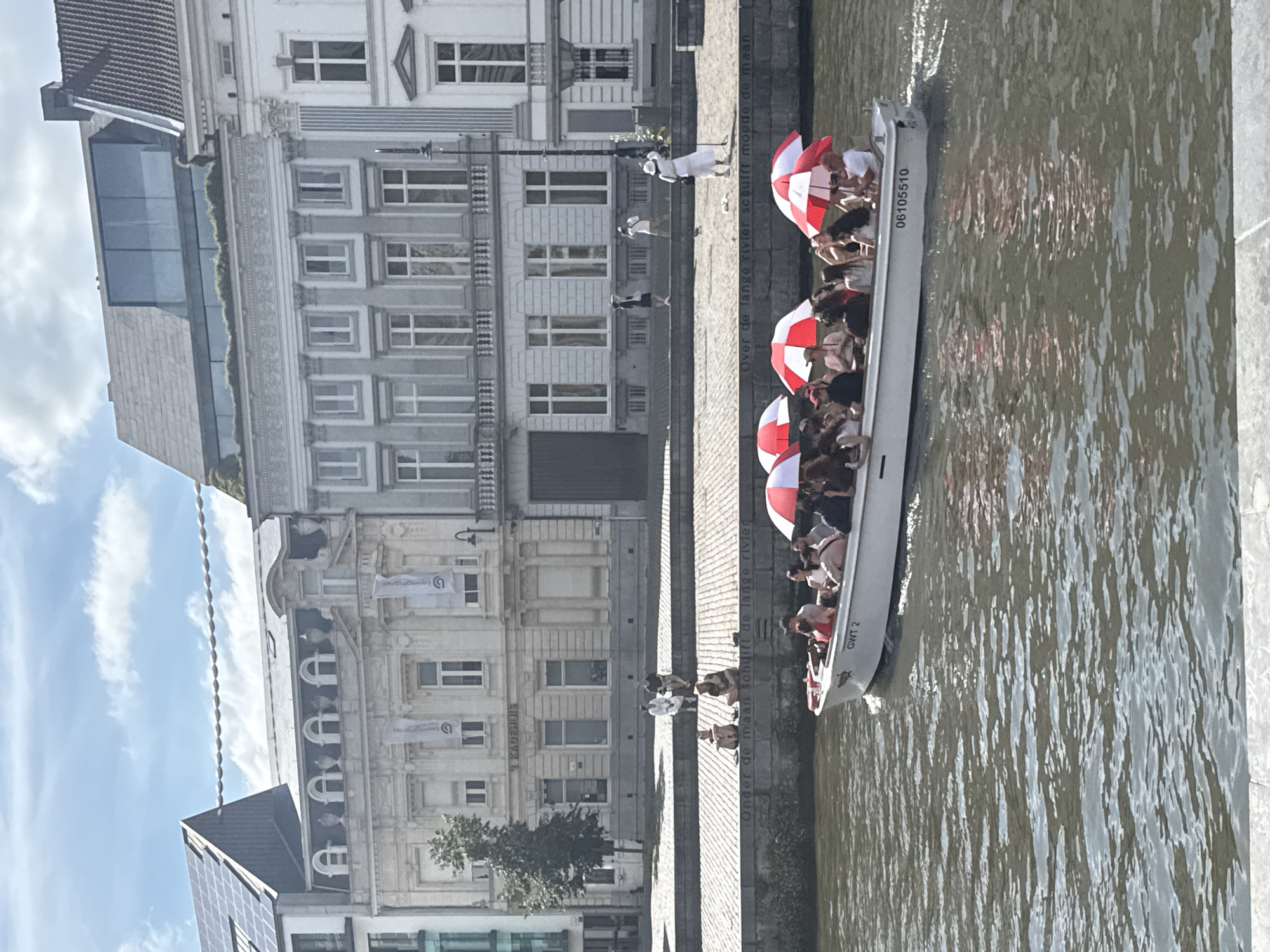 The image captures a bright, sunny day in Ghent, Belgium, focused on a canal and the buildings lining it. In the foreground, a white tourist boat is navigating the water, filled with passengers holding red and white umbrellas. The boat has "GWT 2" and "06105510" visible on its side. The canal water appears choppy with reflections of the sky and surrounding structures.
The middle ground features a stone embankment with a railing. On this embankment, several people are seated, seemingly enjoying the view. One person in a white outfit and hat stands near the railing. The buildings behind the embankment are a mix of architectural styles. The closest building is a light-colored, ornate structure with multiple windows and decorative elements. Further back, there are taller buildings with more modern sections, including one with a glass-fronted upper level and solar panels on its roof. The sky is blue with scattered white clouds. The overall scene suggests a typical tourist activity in a European city.