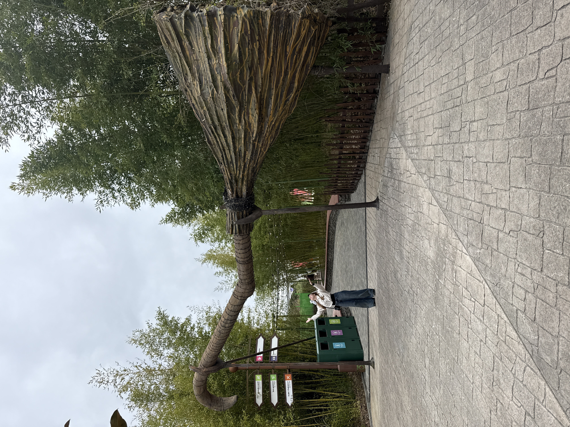 The image depicts an outdoor scene within Sochi Park, Sochi, Russia, featuring a colossal, rustic-themed broomstick sculpture forming an archway over a paved path. The broomstick's curved handle extends from the left, connecting to a multi-directional signpost, while its broad bristle end rests on the right. A single female, positioned beneath the archway on the left side, wears a white shirt, wide-leg dark pants, and a white face mask. She stands with her head tilted back and arms outstretched, appearing to interact playfully with the oversized prop. To her left, the brown signpost displays white arrow-shaped signs with Russian text, pointing towards various attractions like the Dolphinarium and Eco Village. Below the signs, three green waste receptacles with recycling symbols are visible. The pathway consists of irregularly shaped gray and brown flagstones, curving slightly into the background. A low, rustic wooden fence lines the right side of the path. The background is characterized by a dense growth of tall, green bamboo trees. The sky is overcast.