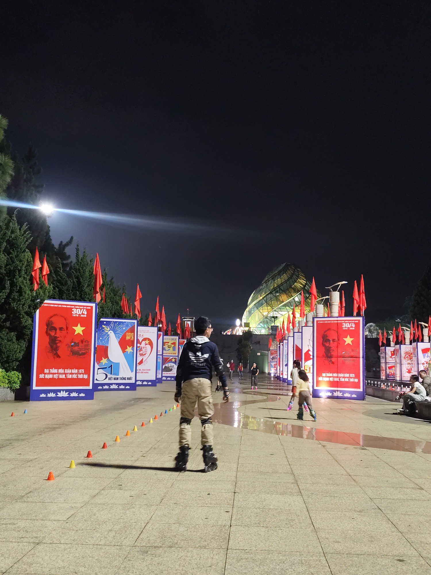 The image shows a night scene on a large paved area in Da Lat, Vietnam. There are several people inline skating. A person is facing away from the camera, with their back visible. Behind them, a line of posters commemorating Vietnamese history are arranged along the walkway, accompanied by red flags. The backdrop features a brightly lit, glass-paneled building with a unique architectural design. Small cones are placed on the ground. The sky is dark. There are trees on the left side of the image. People are sitting on a bench on the right. The pavement appears wet and reflective.