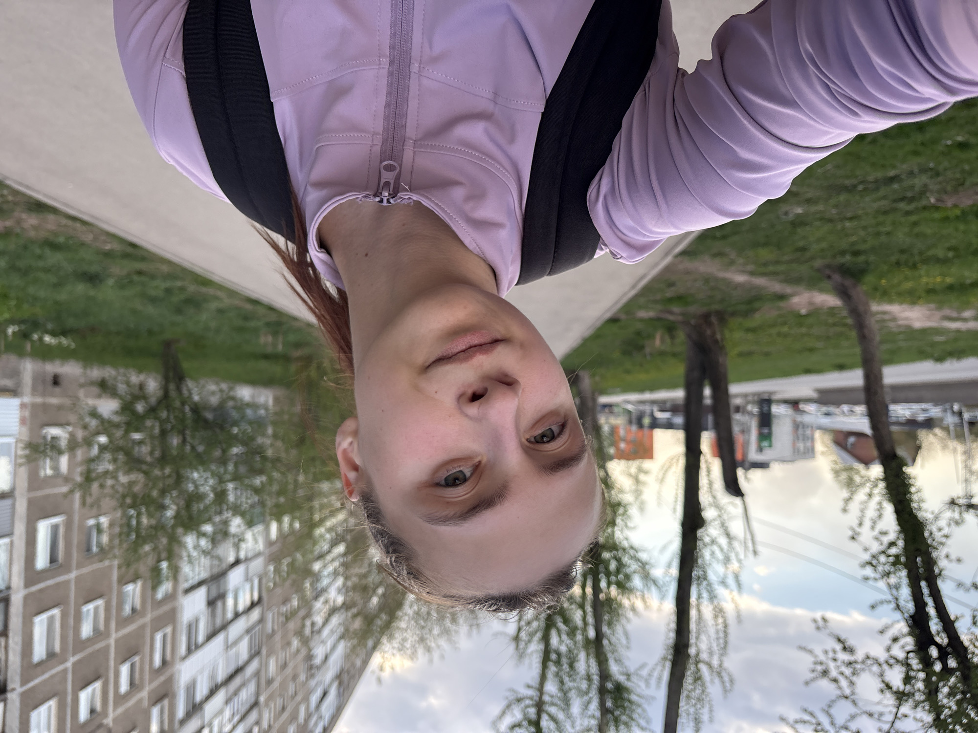 A woman with light skin and light brown hair is facing the camera. She is wearing a light purple zip-up jacket with a black backpack. The image is taken from a low angle, tilting upwards towards the woman.
The background features an urban environment. A grey building with white windows is visible on one side. Trees and sky are also visible. The location appears to be Perm, Russia based on the architecture in the background.