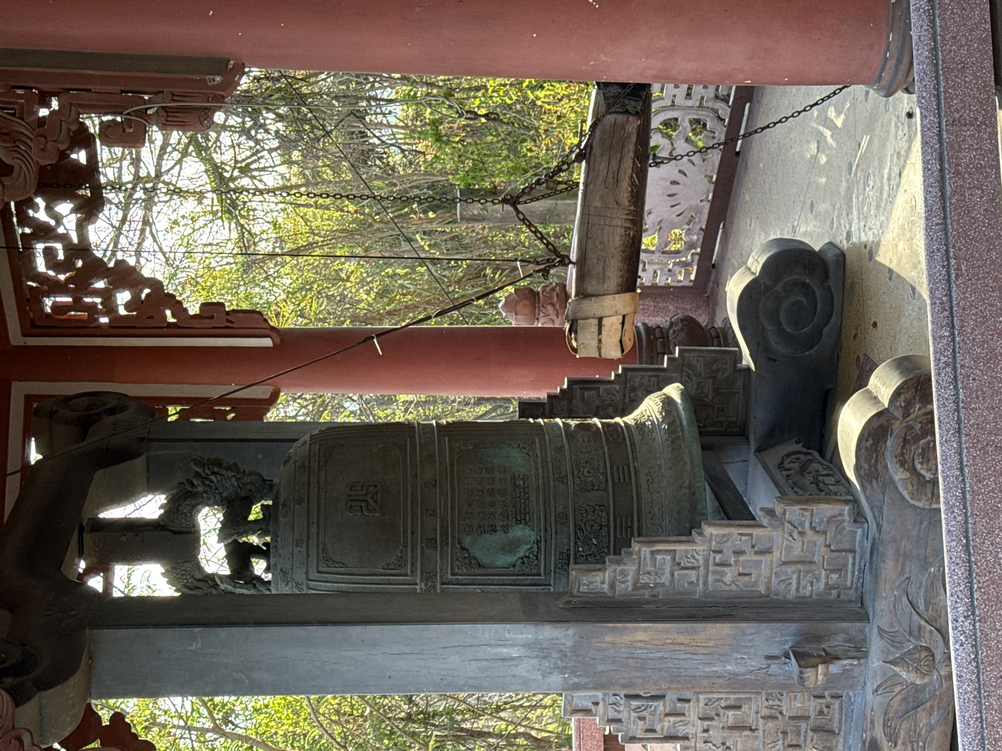 The image depicts a large bronze bell housed within a traditional architectural structure, likely a temple or pavilion, in Tay Nha Trang Ward, Vietnam. The bell is adorned with inscriptions and raised patterns, possibly depicting historical events or religious symbols. It is suspended from a wooden beam by chains. The surrounding structure features intricate carvings, ornate decorations, and traditional architectural elements. The environment is filled with trees and foliage, visible through openings in the structure. The scene is illuminated by natural daylight, casting shadows and highlighting the details of the objects.