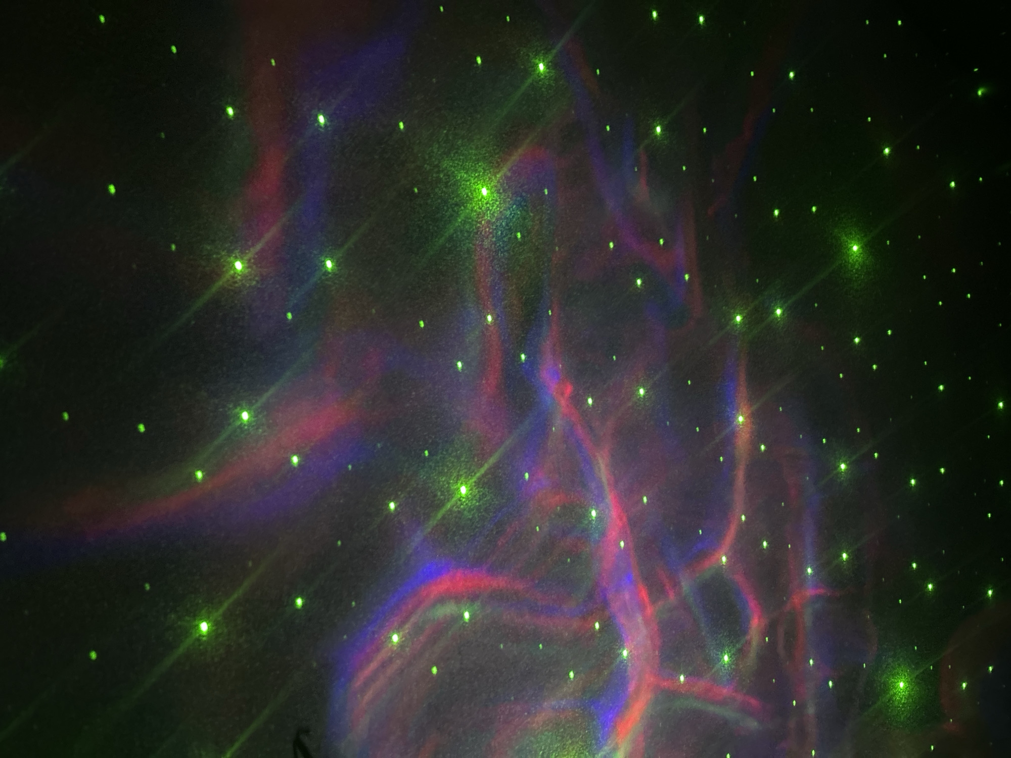 The image shows a dark scene with numerous small, bright green dots scattered across the frame, resembling stars. In the central and left portions, there are blurred patterns of red, blue, and purple light, vaguely resembling nebulas or abstract smoke. The overall impression is that of a simulated night sky or space scene. It is impossible to determine the location, Chaikovsky, Russia, based on the information provided in the image.