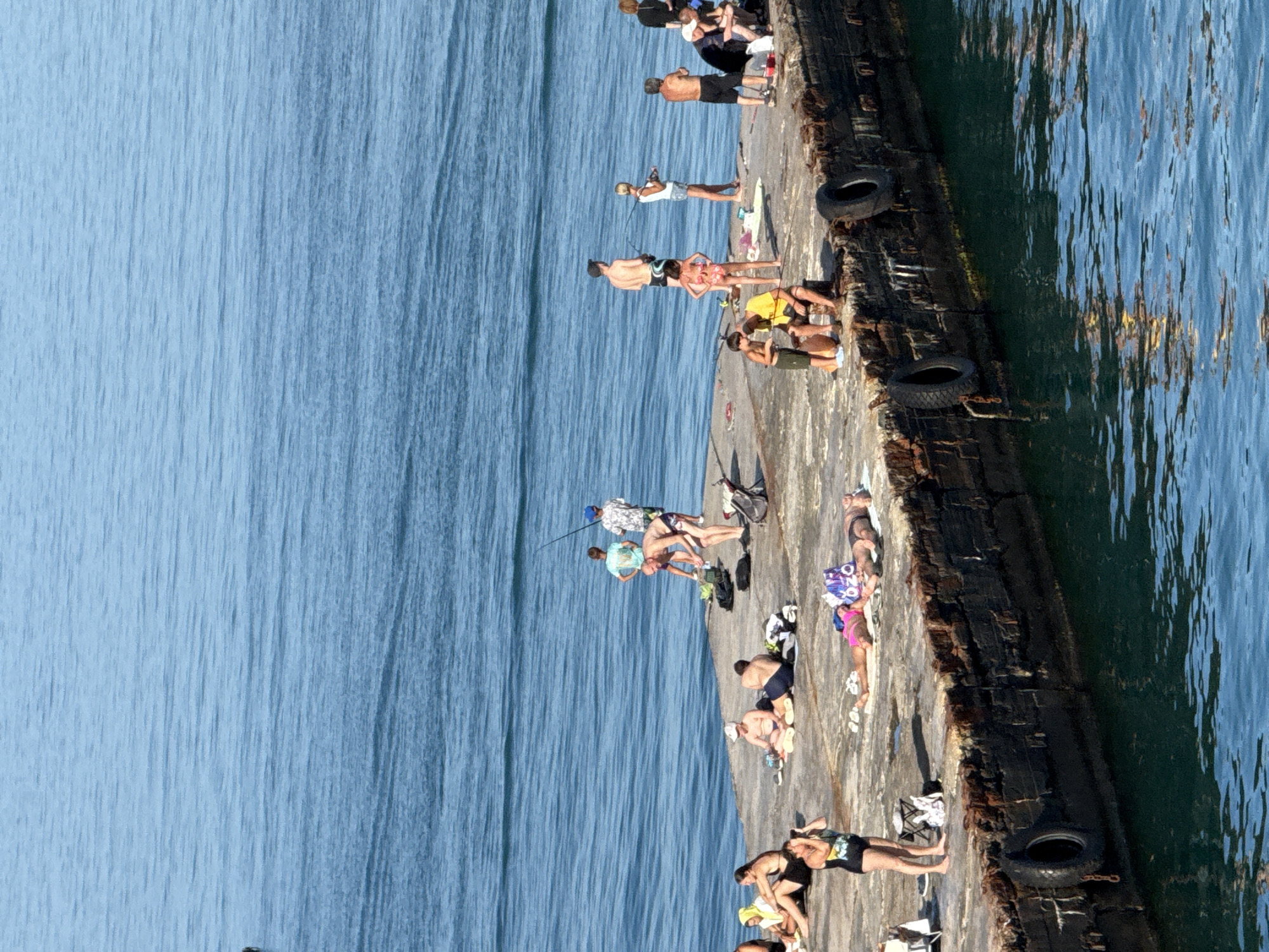 The image depicts a concrete pier extending into a body of blue water, likely the Black Sea. A number of people are present on the pier, engaged in various activities. Several individuals are sunbathing on towels and mats, while others appear to be fishing with rods. Some people are standing, others are sitting or lying down. The pier itself is weathered and appears to be made of concrete blocks, with old tires affixed to the side as fenders. The water is calm, with visible ripples. The scene suggests a leisure activity on a sunny day. Based on the context provided that this image is from Partenit, Ukraine, this is likely a pier in the town.