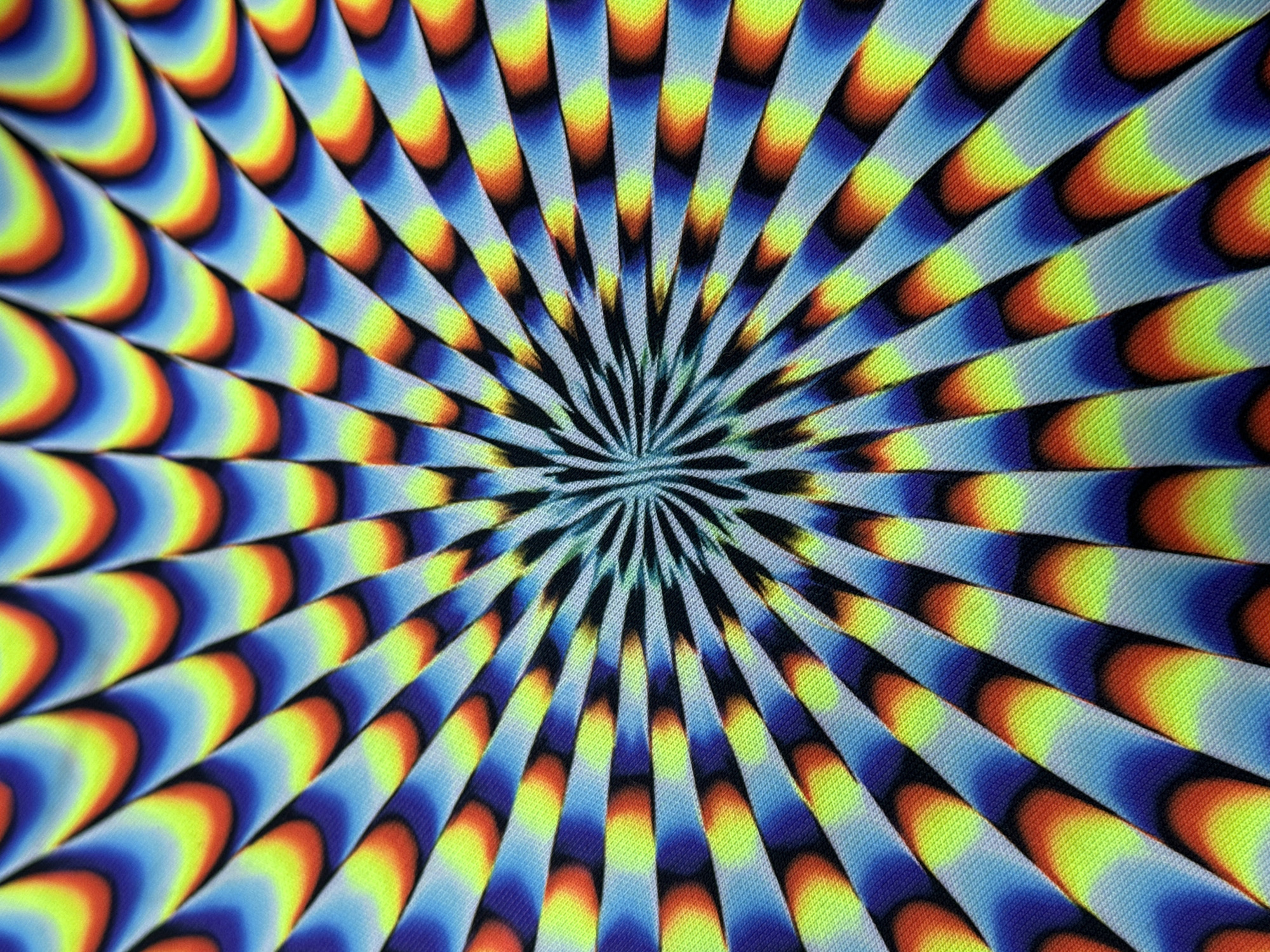 This image is a close-up, abstract shot of a fabric with a radiating, psychedelic pattern. The pattern consists of multiple wedge-shaped segments that converge towards the center. Each segment is filled with a gradient of vibrant colors, including bright yellow, orange, red, and various shades of blue and purple. The texture of the fabric is visible, showing a fine weave pattern. There are no people, identifiable objects, or any indication of a specific location, including Antalya, Turkey. The scene is entirely abstract and decorative.