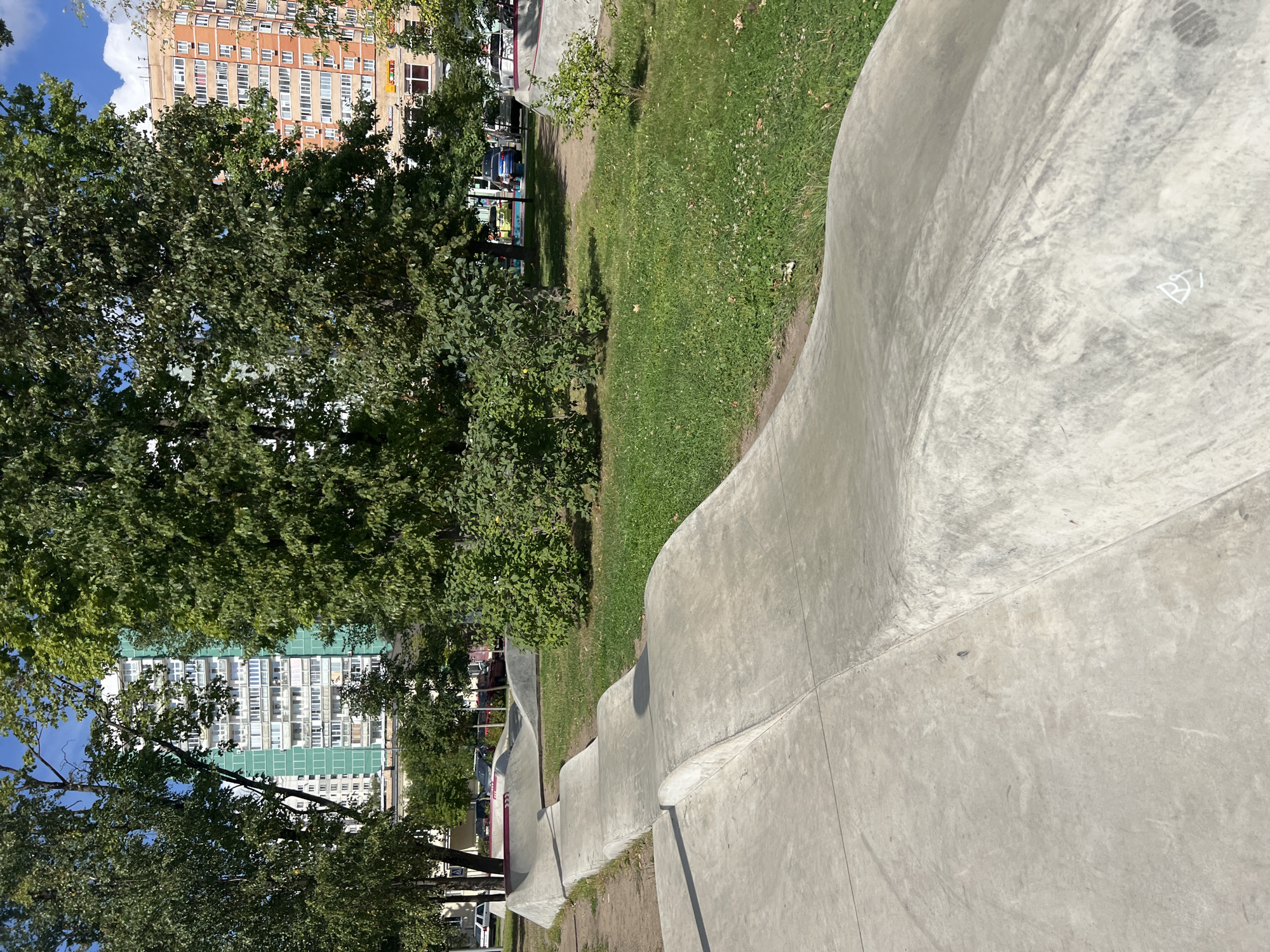 The image depicts an outdoor urban scene in Moscow, Russia, featuring a concrete pump track in the foreground. The pump track consists of a series of smooth, undulating concrete mounds and curves, designed for sports such as skateboarding, BMX biking, and scootering. The surface is light gray and shows signs of wear.
Behind the pump track, to the left, is a large, multi-story residential building painted in shades of green and white. Numerous windows and balconies are visible on its facade. Further left, several tall, leafy trees partially obscure the view of the building.
To the right of the pump track, there is a grassy area with scattered trees and bushes. In the background, another taller residential building, primarily orange and white with many windows, is visible, partially obscured by trees. Several cars are parked on a street or lot in the middle ground. The sky above is blue with scattered white clouds, indicating fair weather. The overall setting suggests a public park or recreational area within a residential neighborhood.