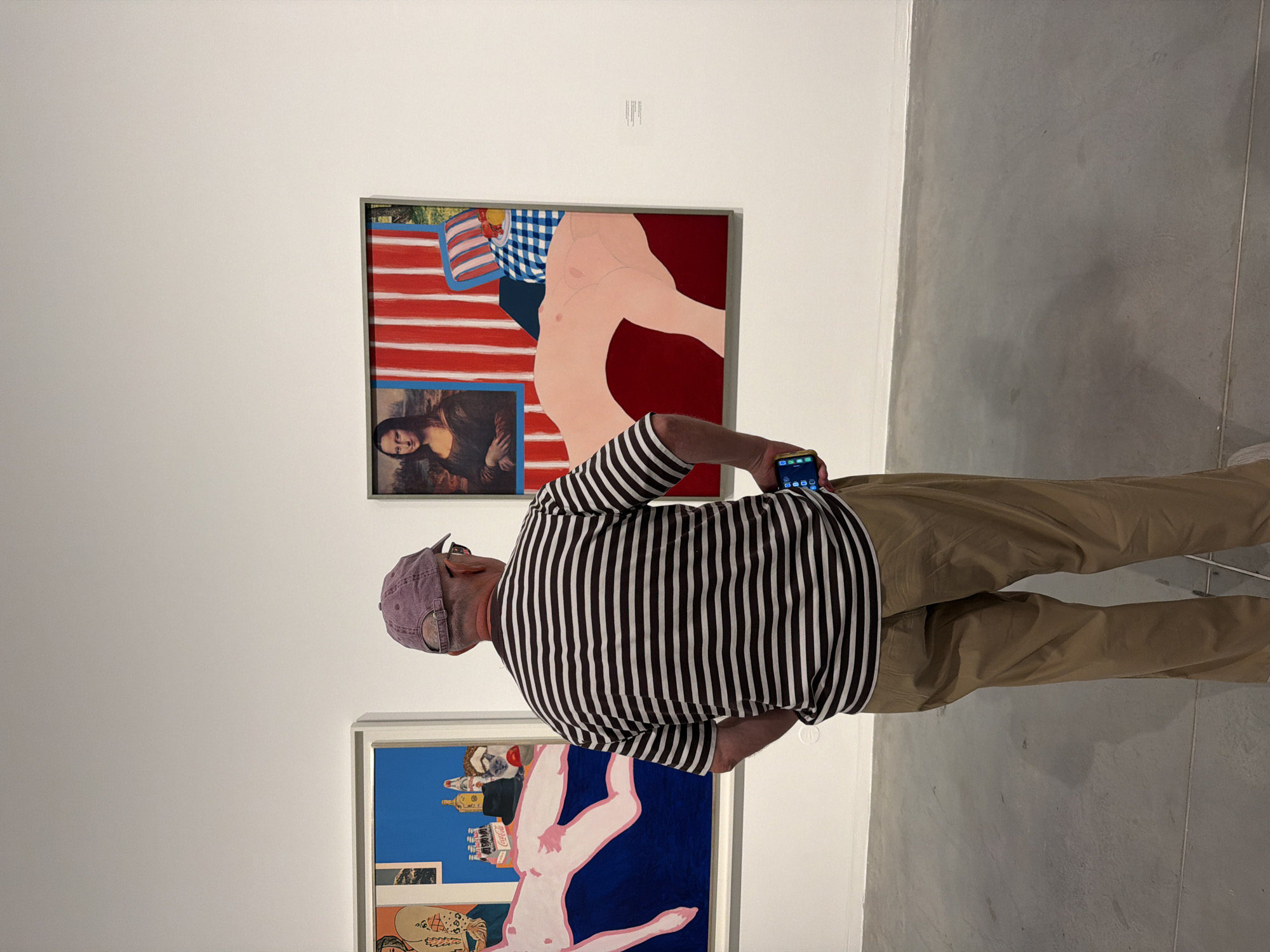 A man wearing a purple baseball cap, a black and white striped t-shirt, and khaki pants stands with his back to the camera, viewing artwork in a gallery. He holds a smartphone in his right hand, with the screen visible. He is facing two large paintings on a white wall. The painting on the left depicts a nude figure lying down, with bottles and a cat. The painting on the right features a striped background, a reclining nude figure, a checkered pillow, and a framed image of the Mona Lisa. The floor is concrete. This scene appears to be in the Tel Aviv Museum of Art, Israel.