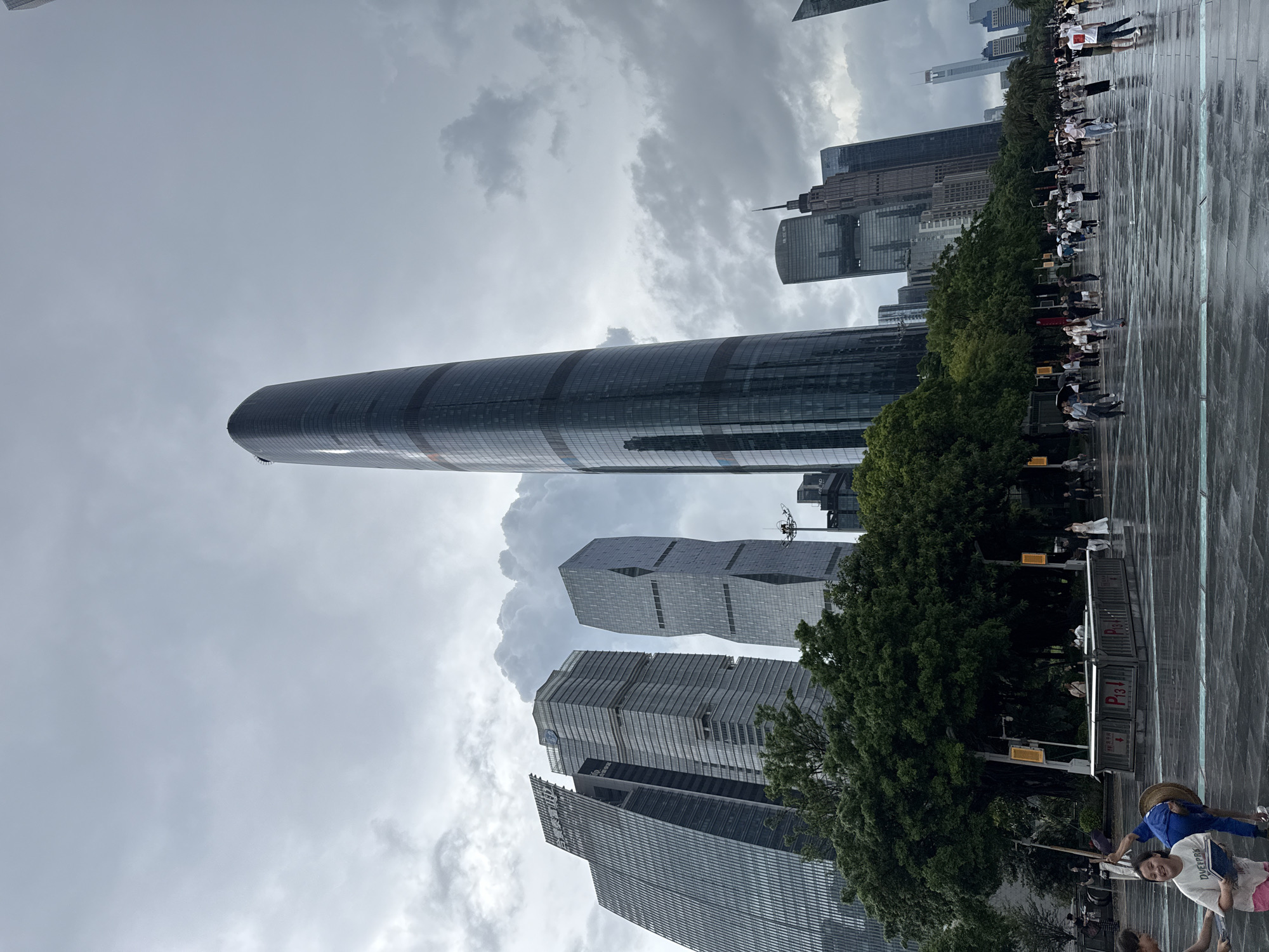The image depicts a wide, wet plaza in front of several modern skyscrapers in Tianhe District, China, under a cloudy sky. The plaza surface, likely made of dark stone or concrete, reflects the overcast sky and the surrounding architecture. Several people are present, walking and interacting in the open space. In the foreground, a group of children are on the wet ground, with one child in a white t-shirt and pink shorts looking towards the camera and holding a blue object. Another child, wearing a blue outfit and a straw hat, is in a dynamic pose, bending over with one leg raised. To the left, another child is partially visible.
Behind the people and the plaza, a line of lush green trees provides a natural boundary before the towering buildings begin. The architecture is predominantly glass and steel, with several distinct skyscrapers of varying heights and designs. The most prominent building on the right is a tall, slender, cylindrical skyscraper with a dark glass facade. To its left are other modern office buildings with angular designs and reflective surfaces. In the background, more skyscrapers extend towards the horizon, some with distinct architectural features like spires. The overall atmosphere suggests a public urban space, possibly a park or a thoroughfare, experiencing overcast weather, indicated by the dark, diffused lighting and the wet ground. Several yellow light poles are visible along the tree line.