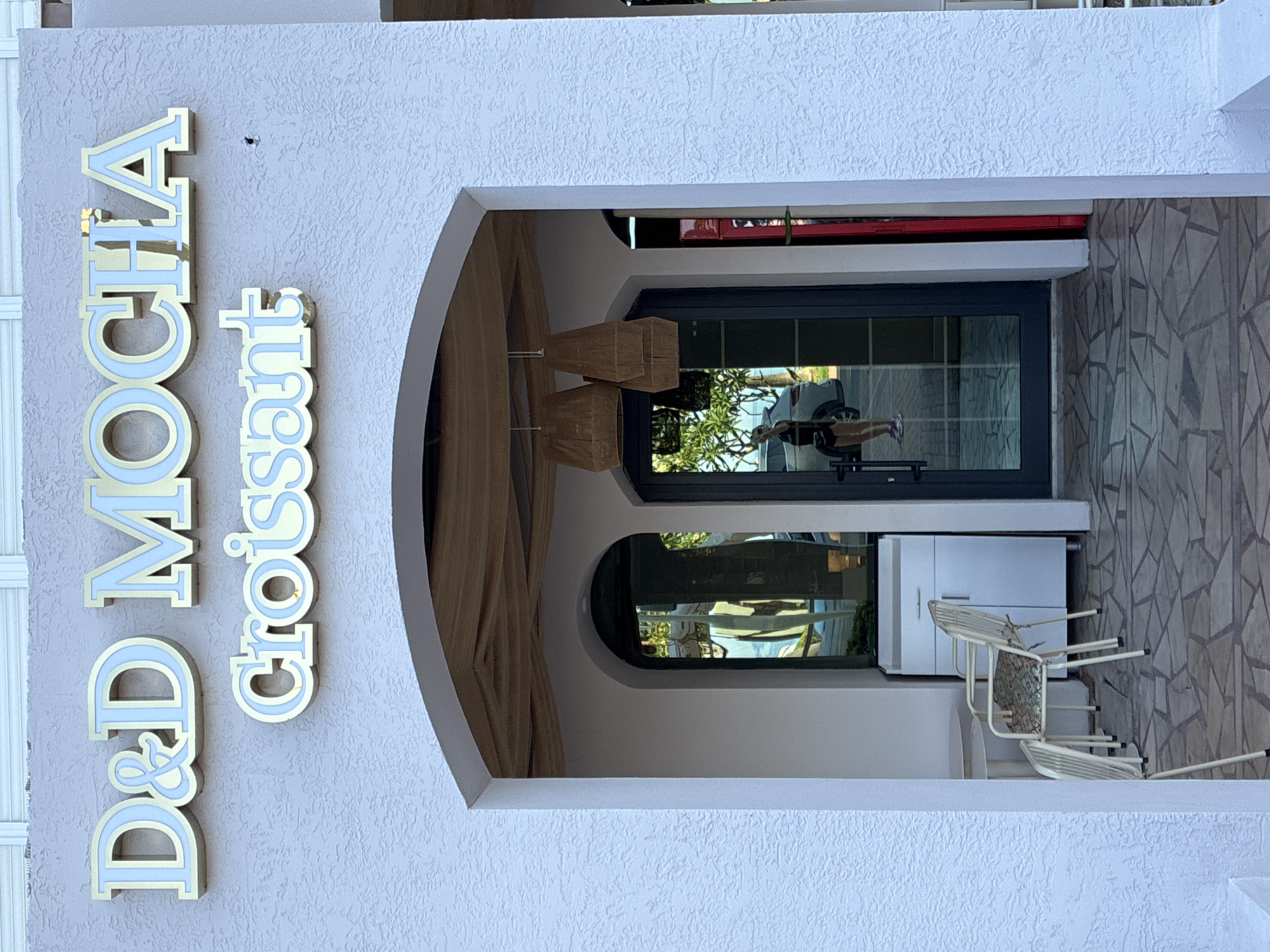 The image shows the facade of a shop named "D&D MOCHA Croissant" in Alanya, Turkey. The shop has a light blue textured exterior wall with gold and light blue lettering. An arched entrance leads into the shop, with glass doors and windows reflecting the surroundings. Inside, two wicker lamps hang from the ceiling. To the left of the entrance, there are two white chairs and a round table. A white cabinet is visible to the right of the chairs. The ground is paved with irregularly shaped stones. A person is visible in the reflection of the glass doors, wearing a black top and shorts.