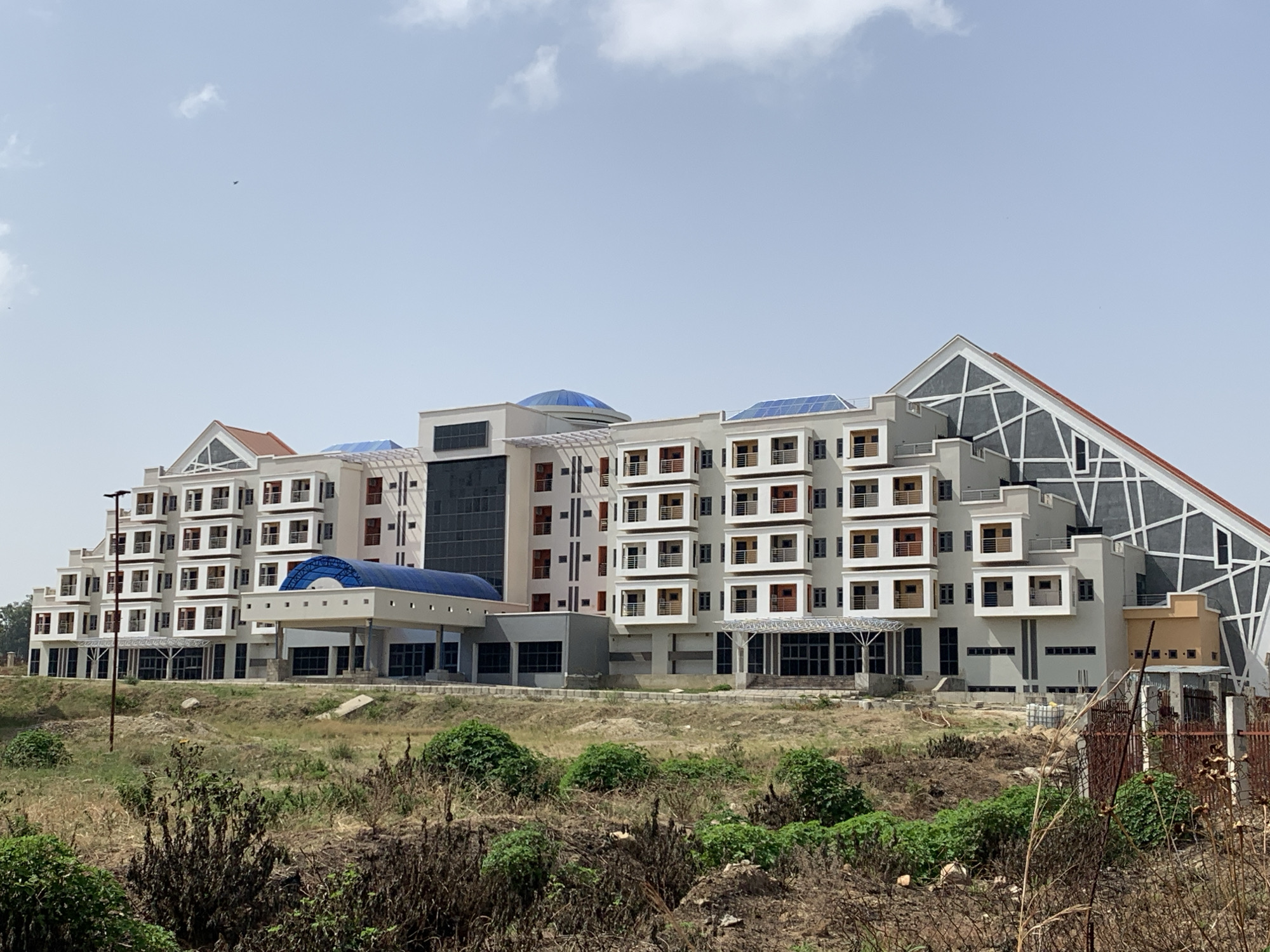 The image depicts a large, multi-story building with a contemporary architectural style. The building is characterized by a mix of white and grey facades, with sections featuring balconies and decorative window frames in muted earth tones. Distinctive elements include a glass-encased, triangular section at one end and a blue-domed structure on the roof.
The foreground is dominated by unkempt vegetation, suggesting an undeveloped or recently disturbed landscape. A rusty metal fence is partially visible on the right side of the frame. The sky is pale blue with scattered clouds, indicating a bright, daytime setting.
The location of the building is unspecified.