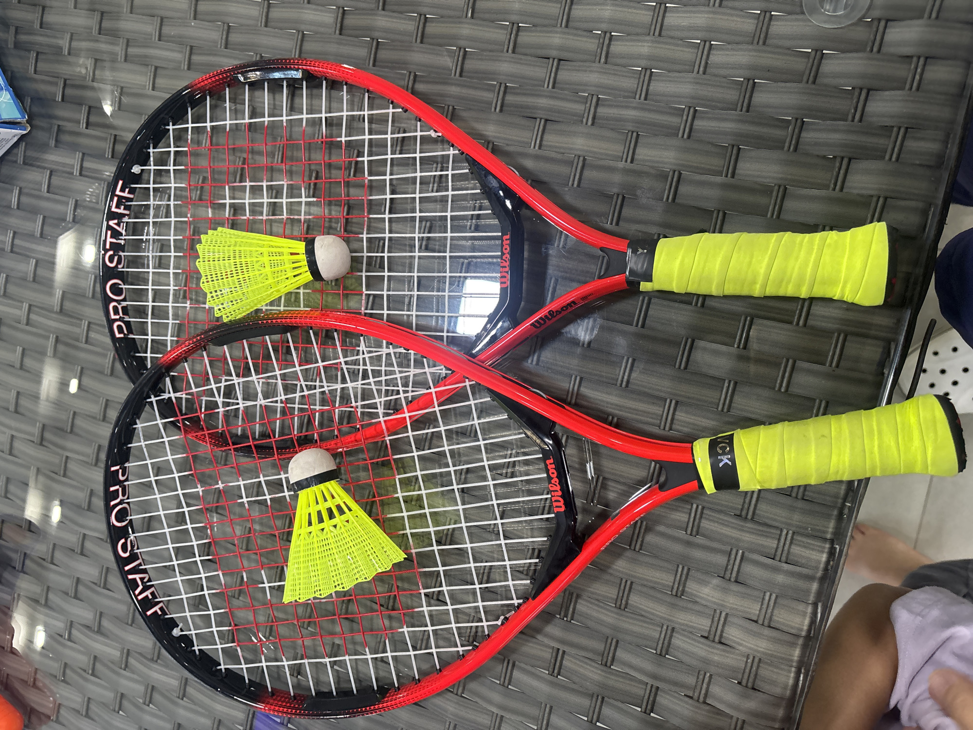 Two red and black badminton rackets with yellow grips and yellow shuttlecocks are laid out on a gray woven table. One racket has the words "PRO STAFF" printed on it. The other racket has "Wilson" printed on the frame and "Wilson" printed on the handle. The table appears to be outdoors. There are also parts of two people visible, one wearing a purple shirt and the other wearing dark blue pants. The scene is likely in Dubai, United Arab Emirates, given the common availability of outdoor tables in residential or recreational areas.