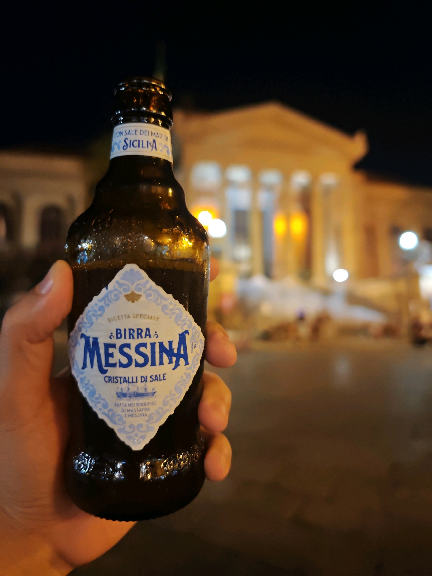 A hand holds a dark glass bottle of Birra Messina beer at night, in front of a brightly lit building with columns, likely a historic structure in Palermo, Italy. The beer bottle has a white label with blue text that reads "BIRRA MESSINA" and "CRISTALLI DI SALE." The background building is illuminated and appears to be a place of significance, possibly a theater or a public institution, with warm light emanating from its windows. The overall scene suggests an evening outing or celebration.